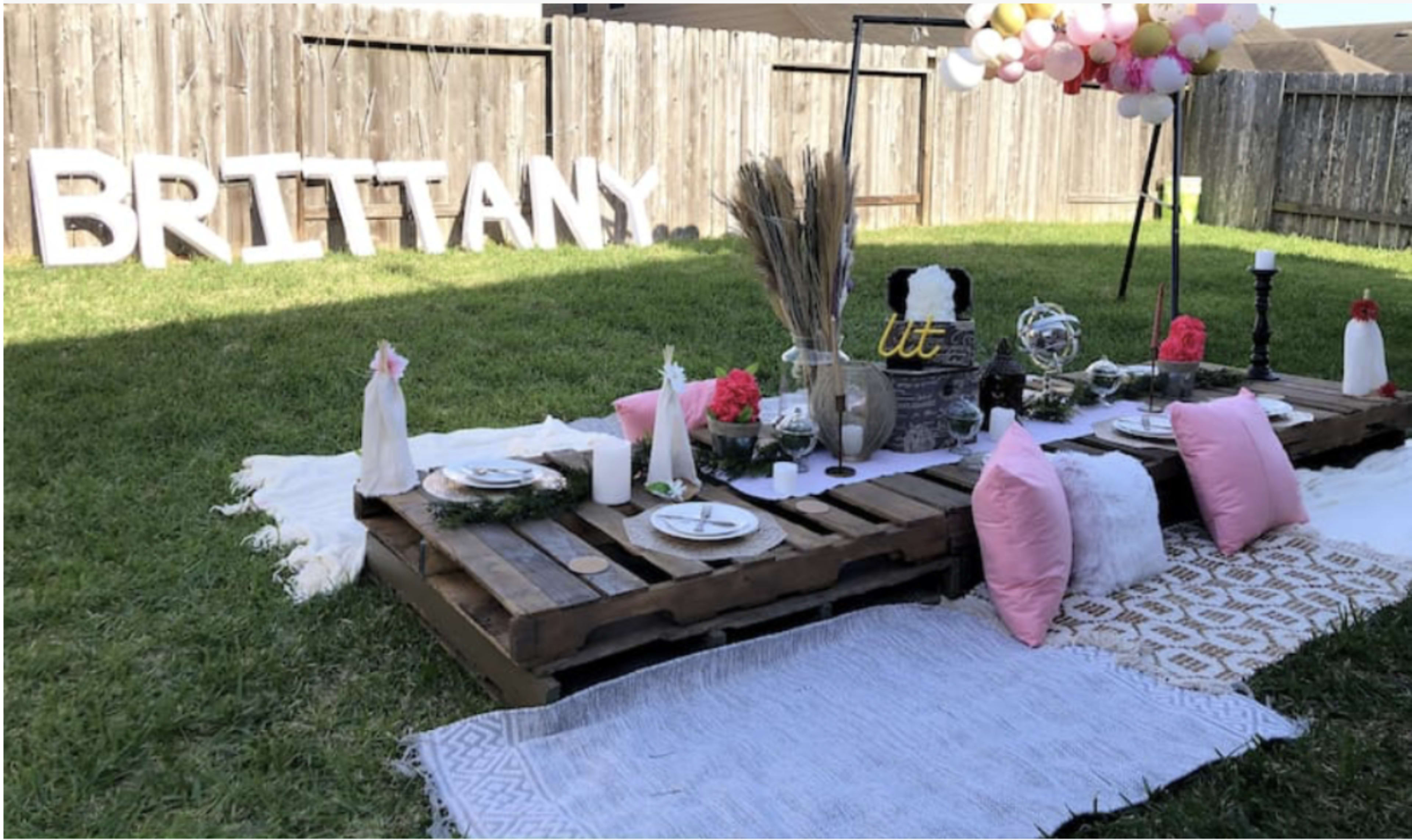 The image shows a decorated outdoor picnic setup on grass, featuring a low wooden pallet table adorned with plates, glasses, and decorative items, alongside large letters spelling "BRITTANY" and a floral arrangement overhead.