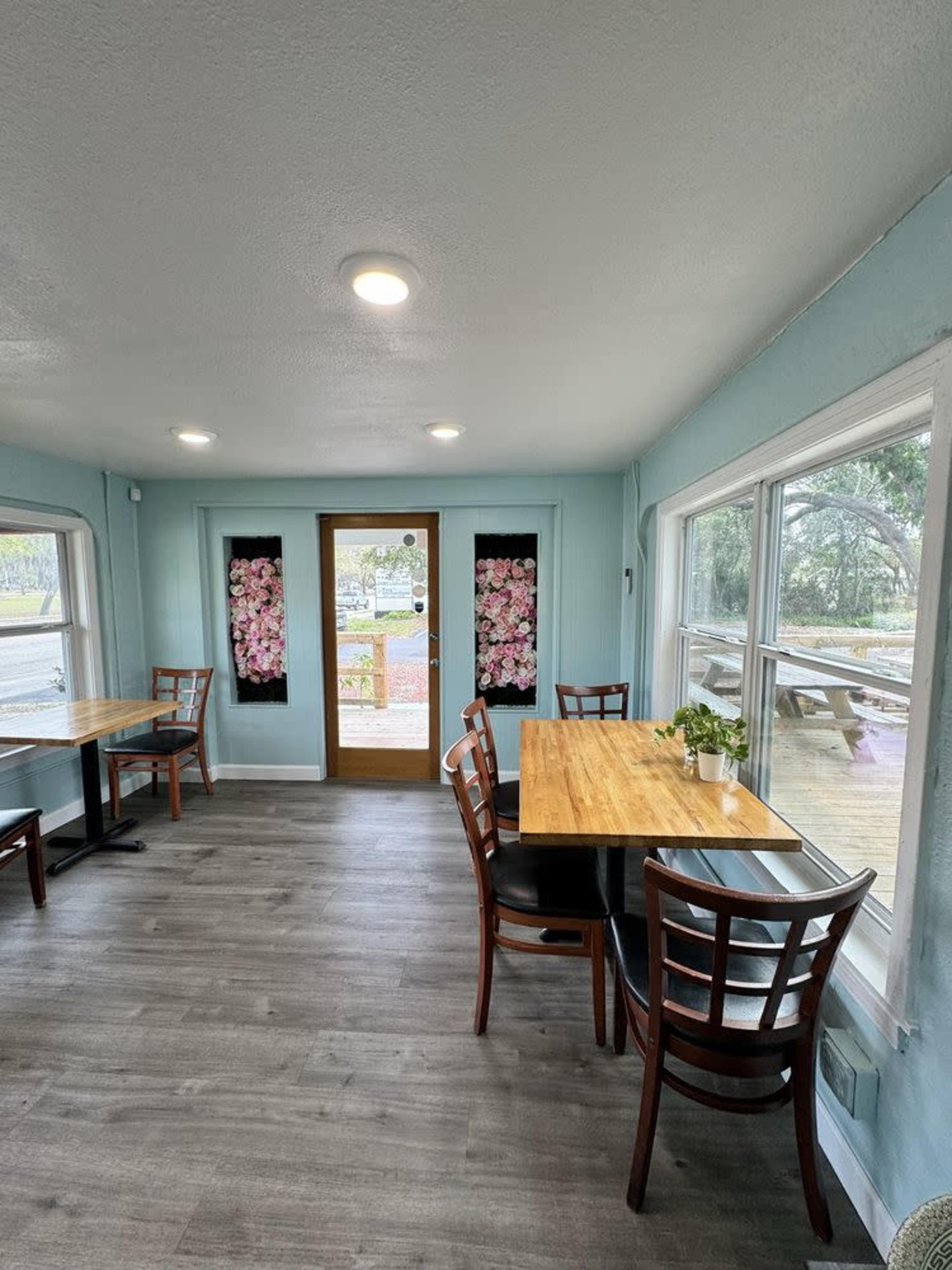 The interior of a small restaurant features wooden tables and chairs, light blue walls, and large windows framed with floral curtains.