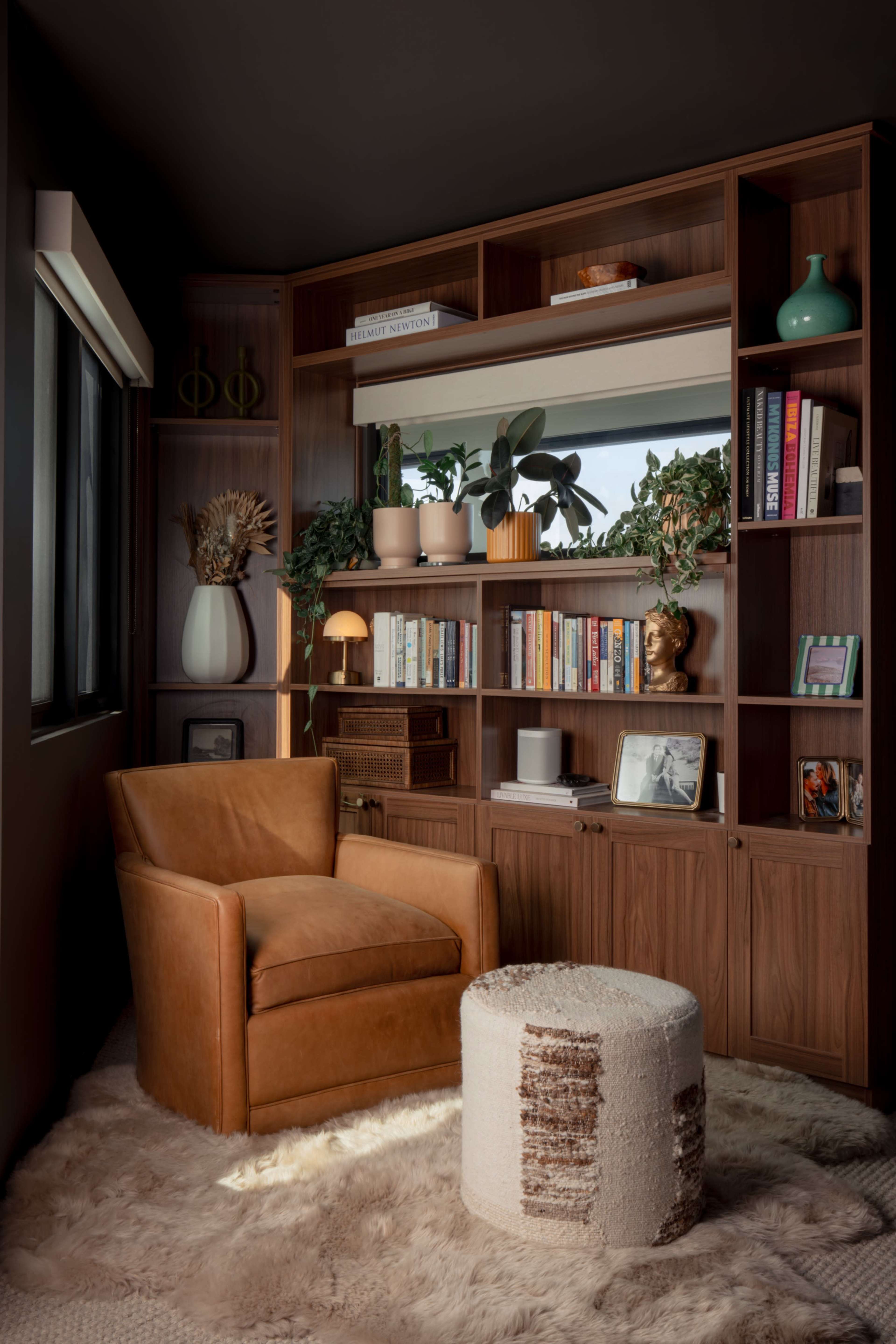 A cozy reading nook features a brown chair and a textured ottoman, surrounded by a wooden bookshelf filled with books and decorative items, against a backdrop of plants and a window.