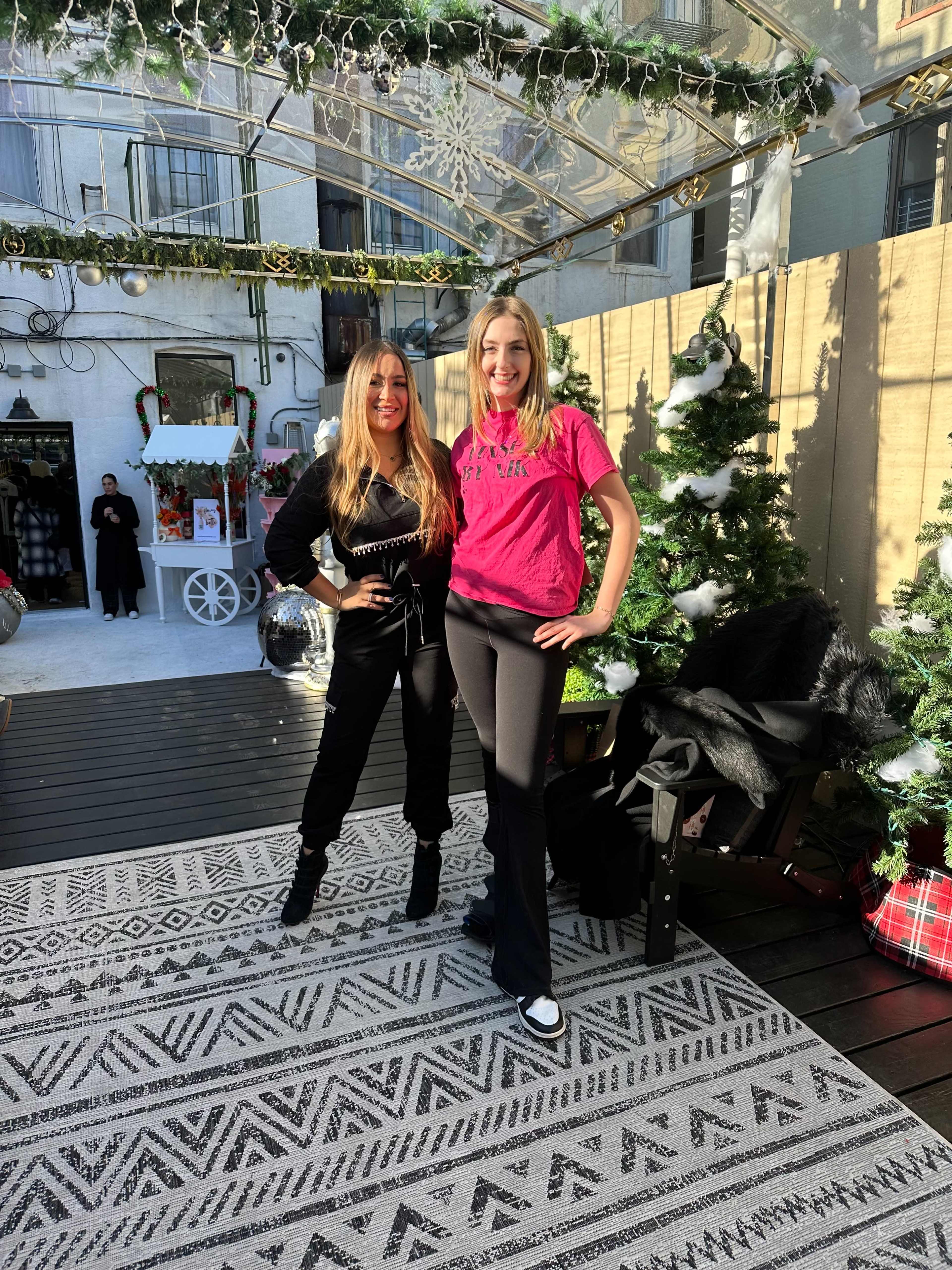Two women pose together in a decorated outdoor space adorned with Christmas trees and holiday decorations.