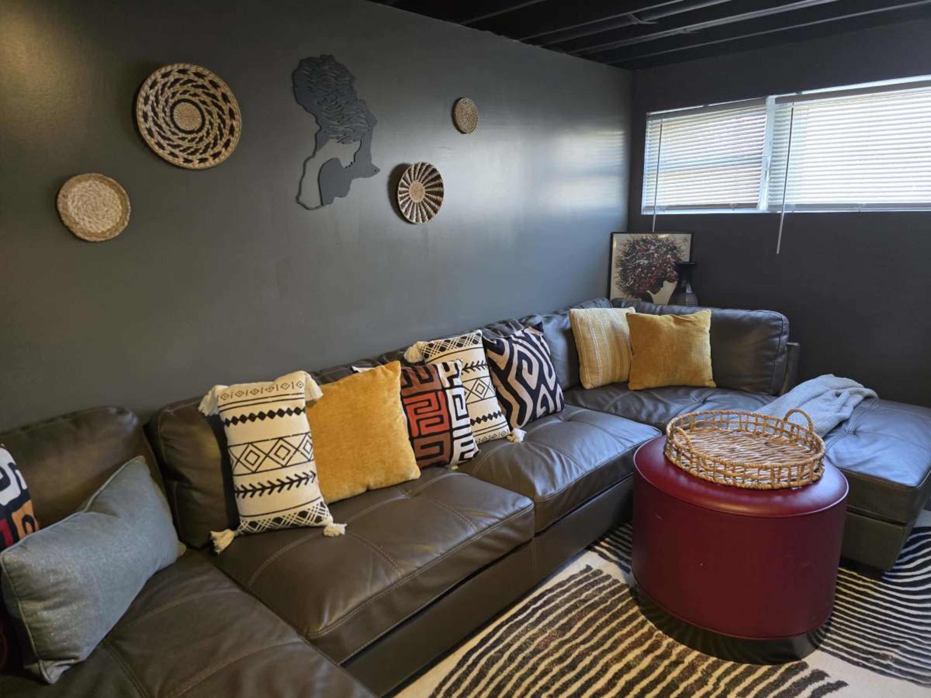 The image shows a cozy living room corner featuring a large sectional sofa with patterned pillows, a round red table, and various decorative wall hangings.