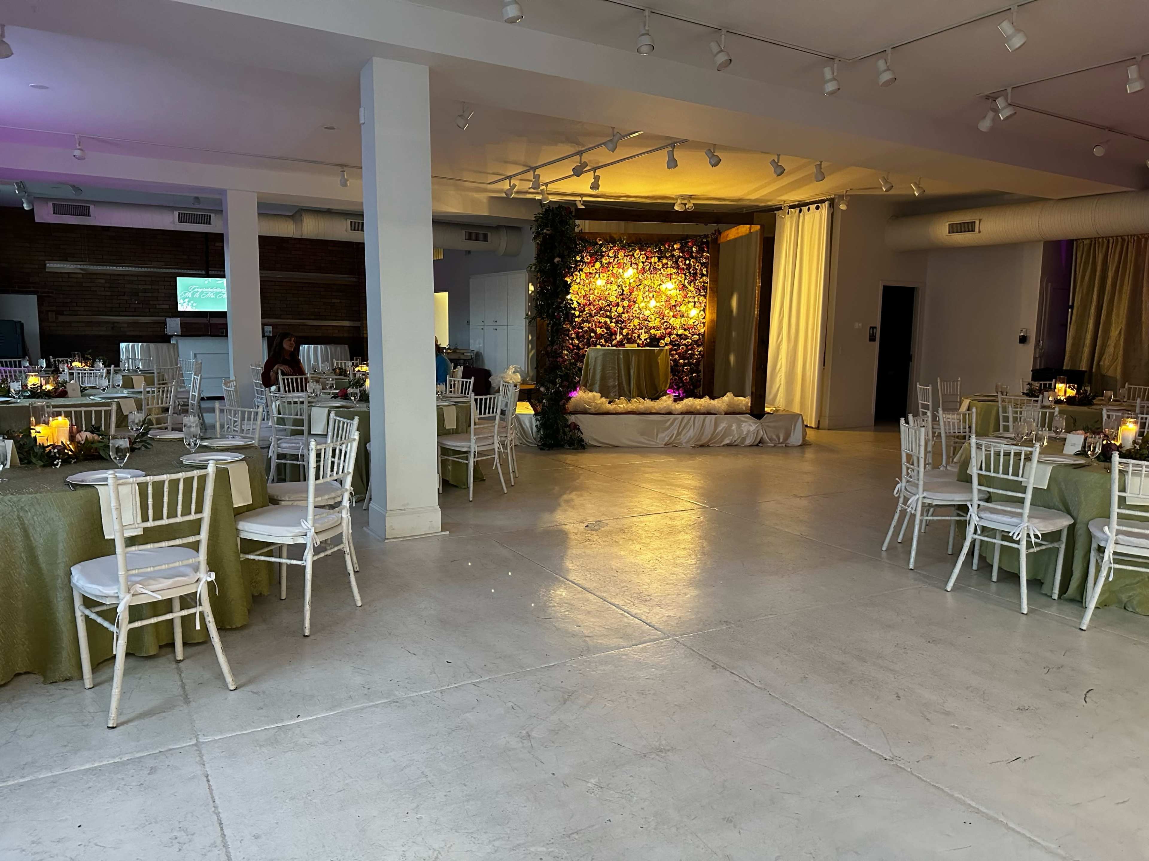 An indoor event space with round tables set for a gathering, a floral backdrop on a stage, and various decorations in muted colors.