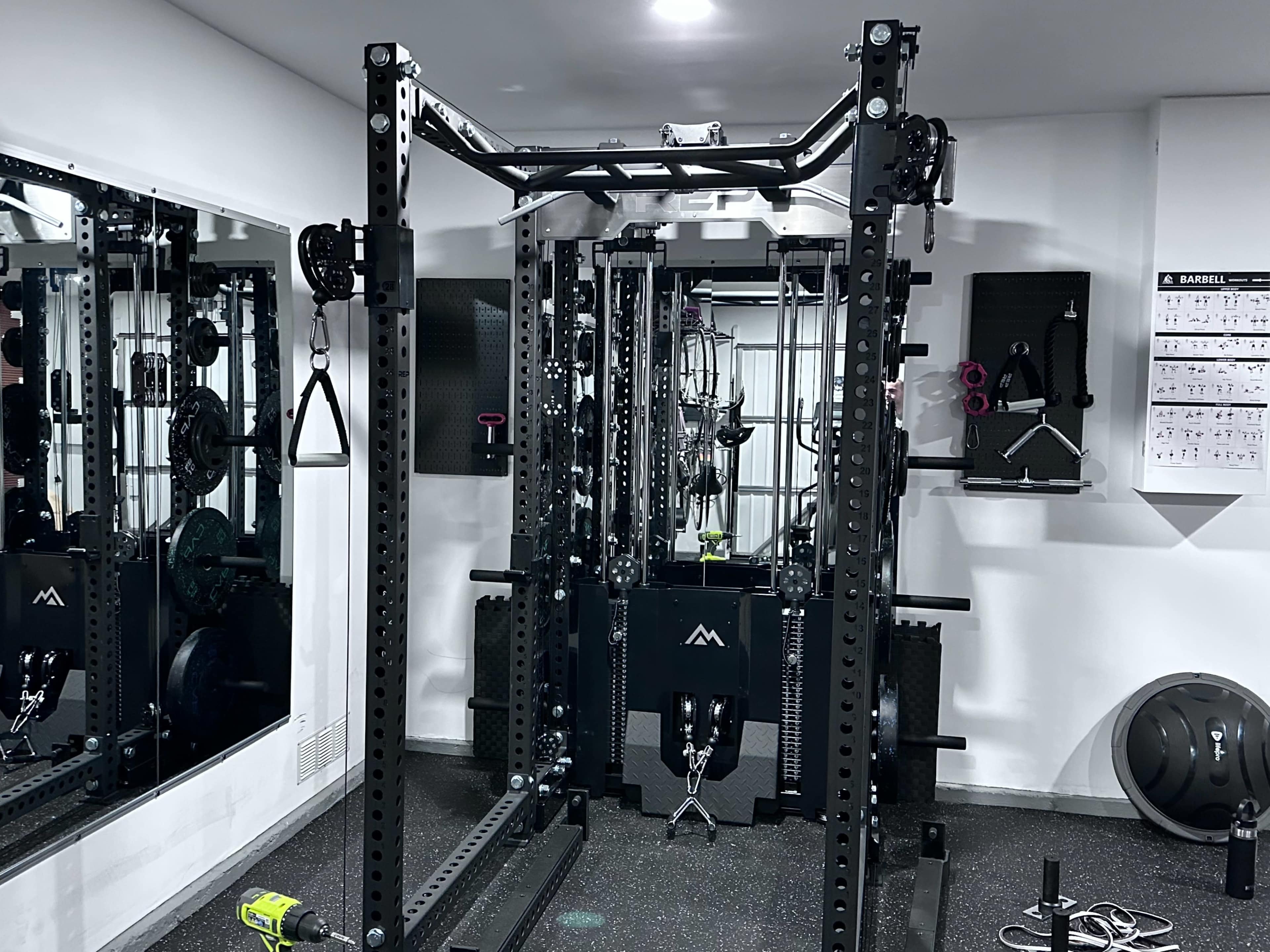 The image shows a well-equipped home gym with a cable machine, pull-up bar, and various weights visible in mirrors and on racks.