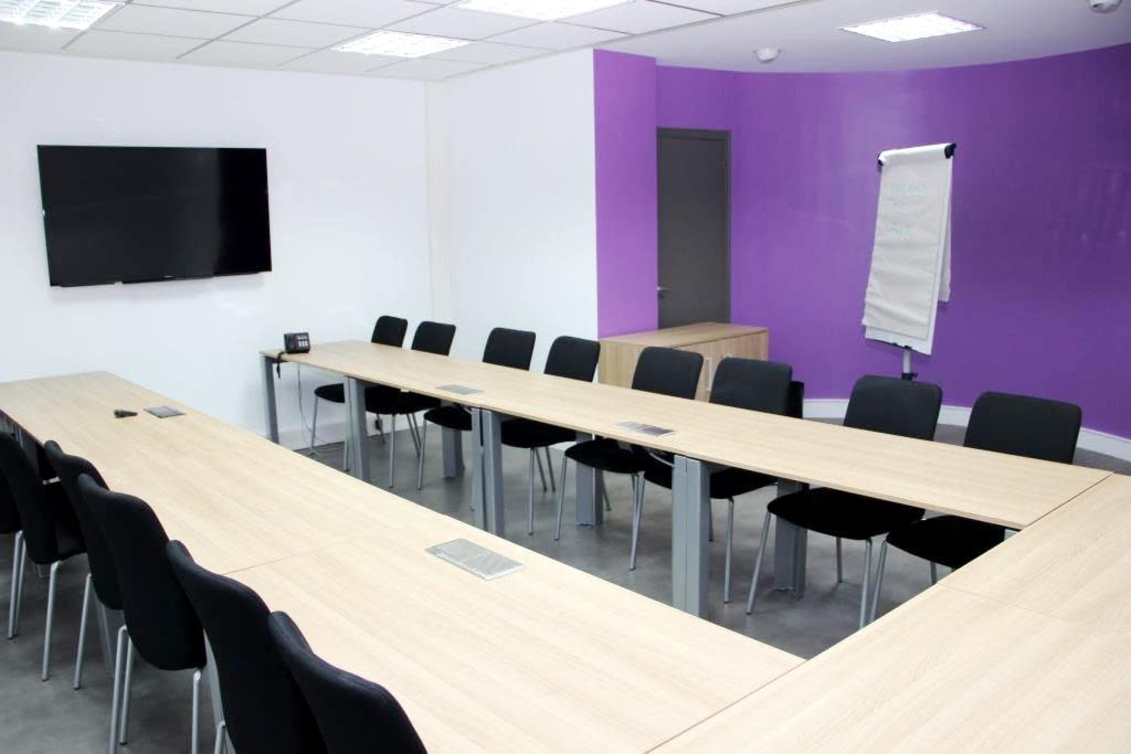 A meeting room features a long wooden table surrounded by black chairs, with a television screen mounted on the wall and a purple accent wall.