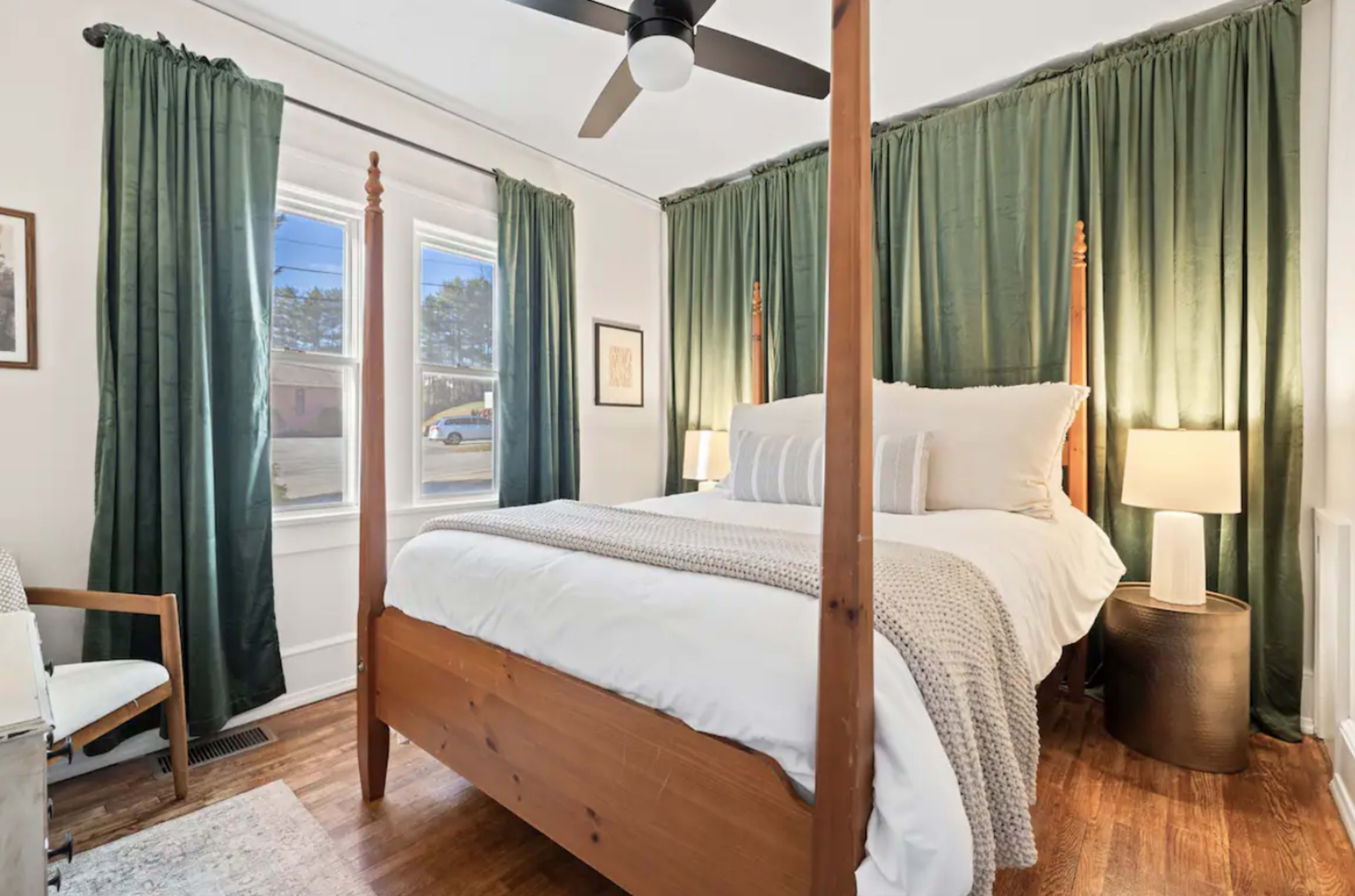 A bedroom with a four-poster bed, green curtains, and a chair next to a lamp on a side table.