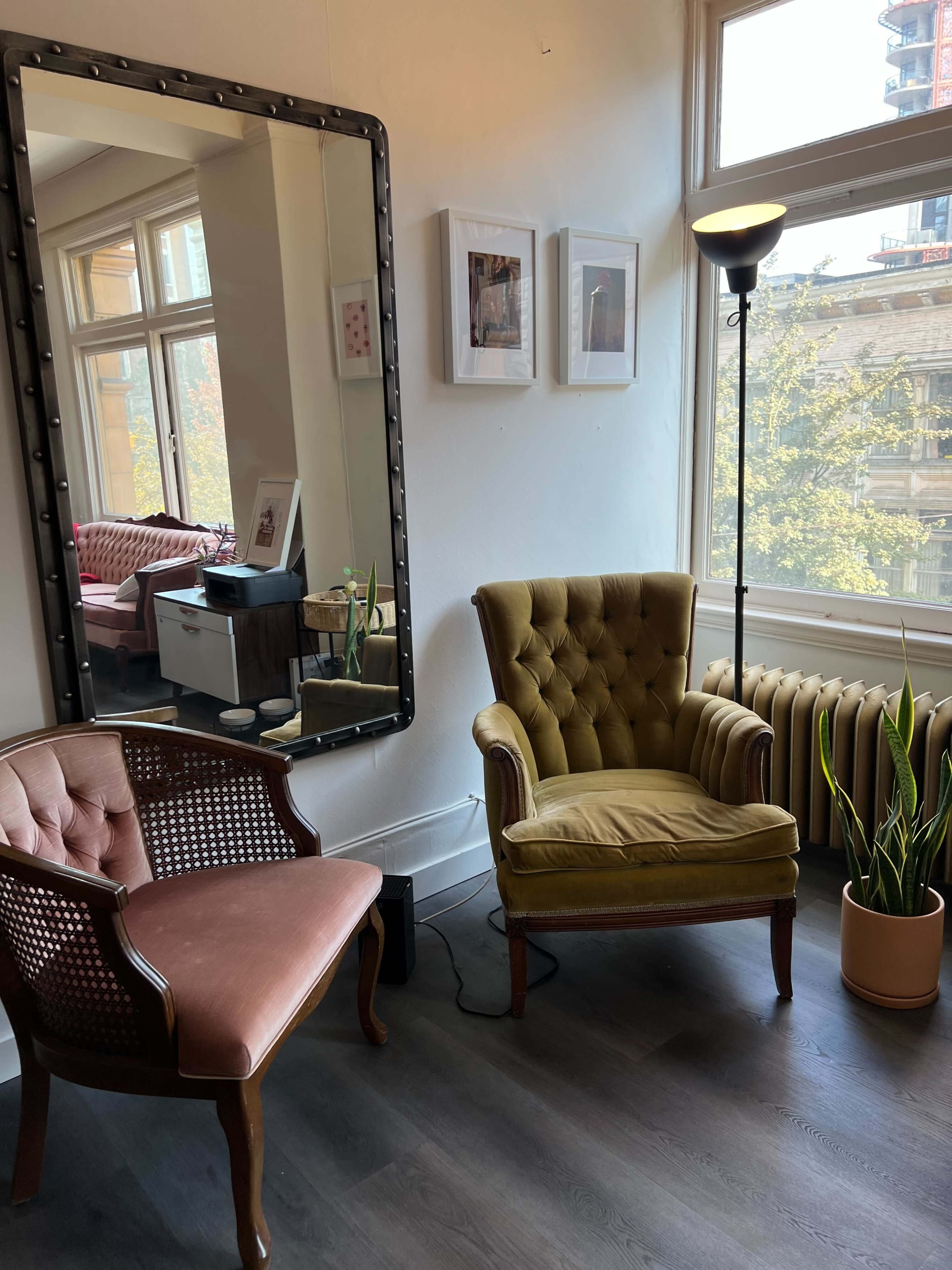 A cozy room with two armchairs, a large mirror, framed pictures on the wall, and a plant beside a floor lamp, all illuminated by natural light from a window.