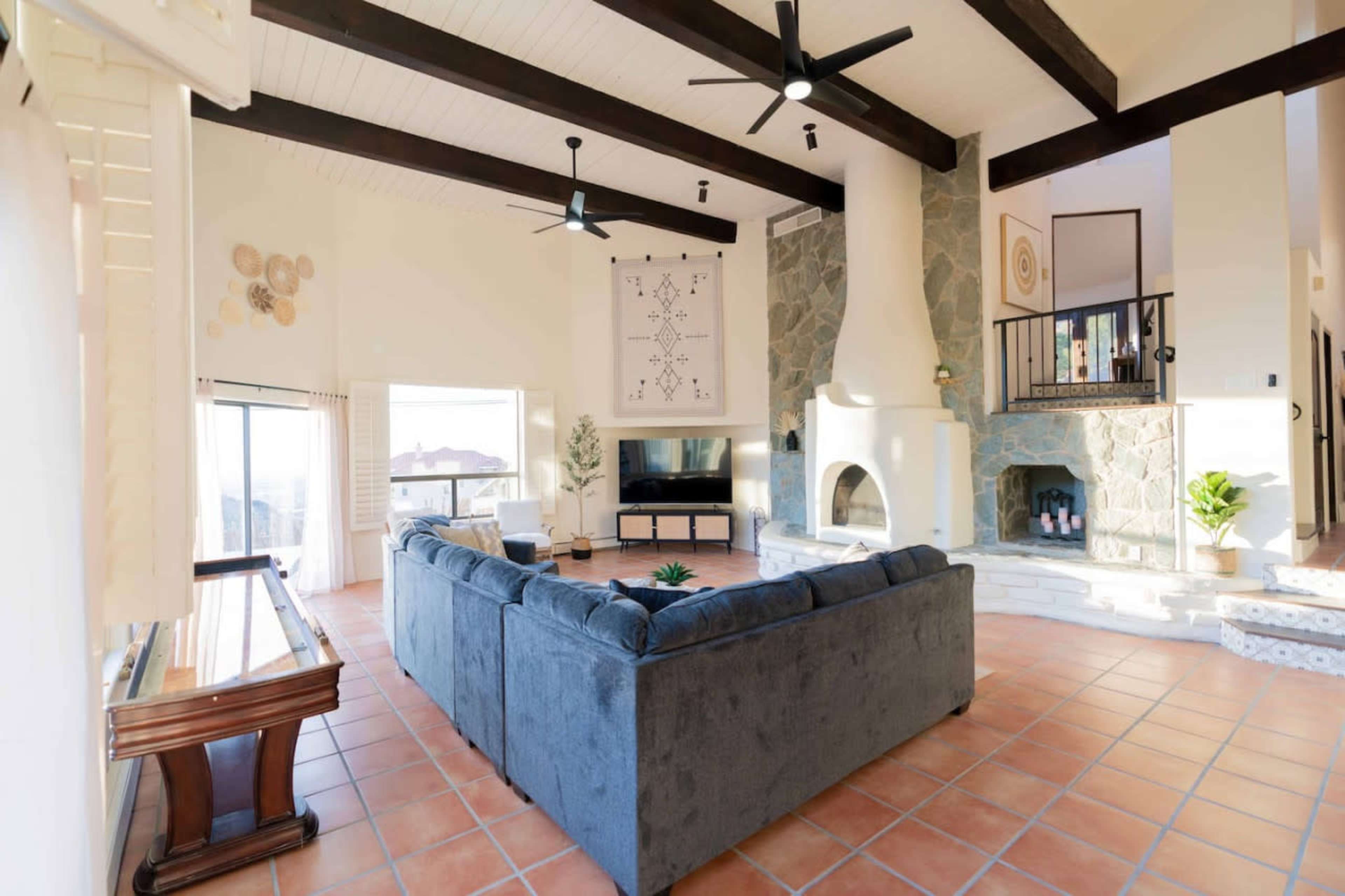 A spacious living room with a high ceiling, featuring a stone fireplace, large windows, and a sectional sofa arranged around a central area.