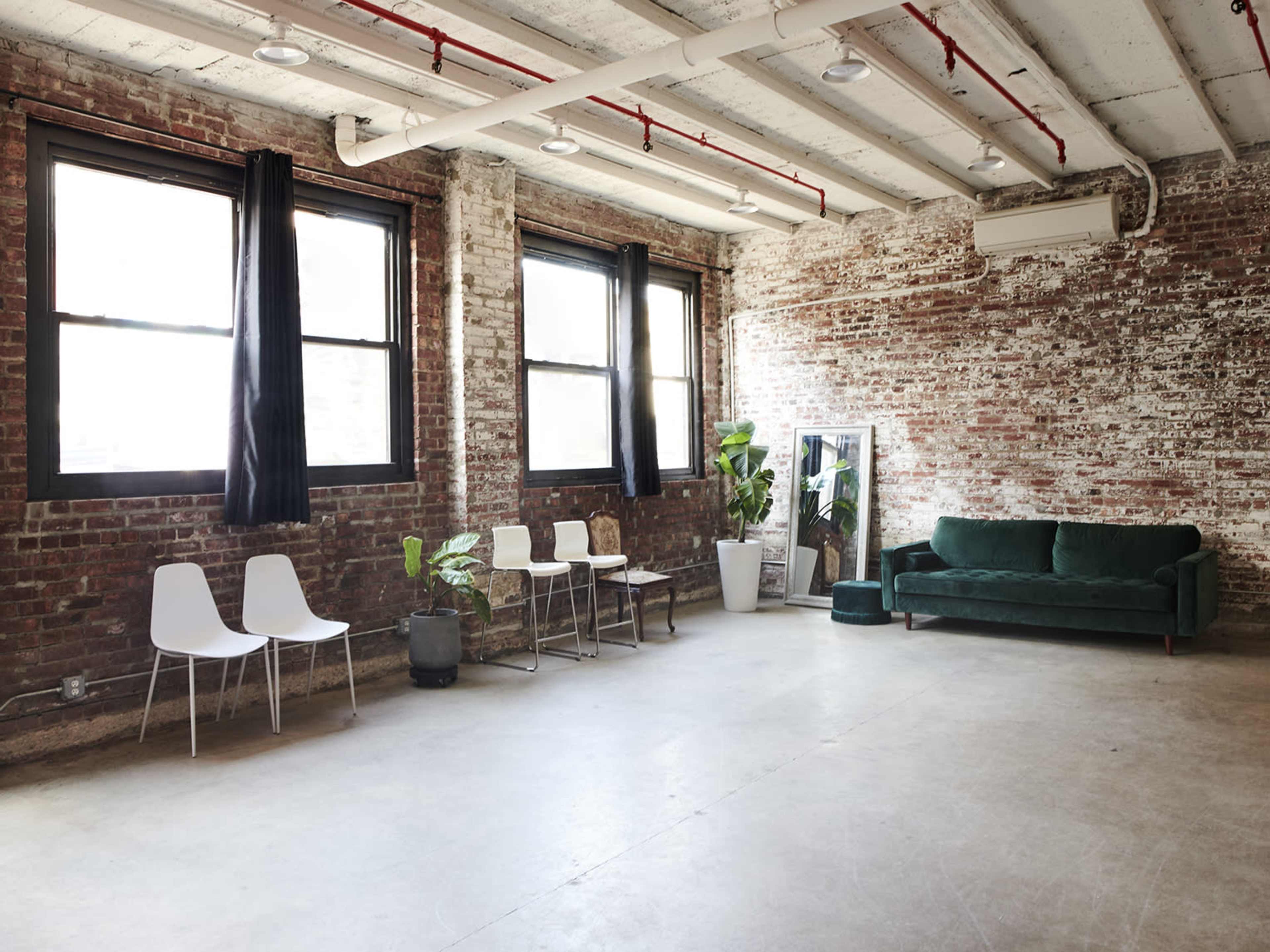 The room features exposed brick walls, large windows, a green sofa, minimalist chairs, and potted plants.