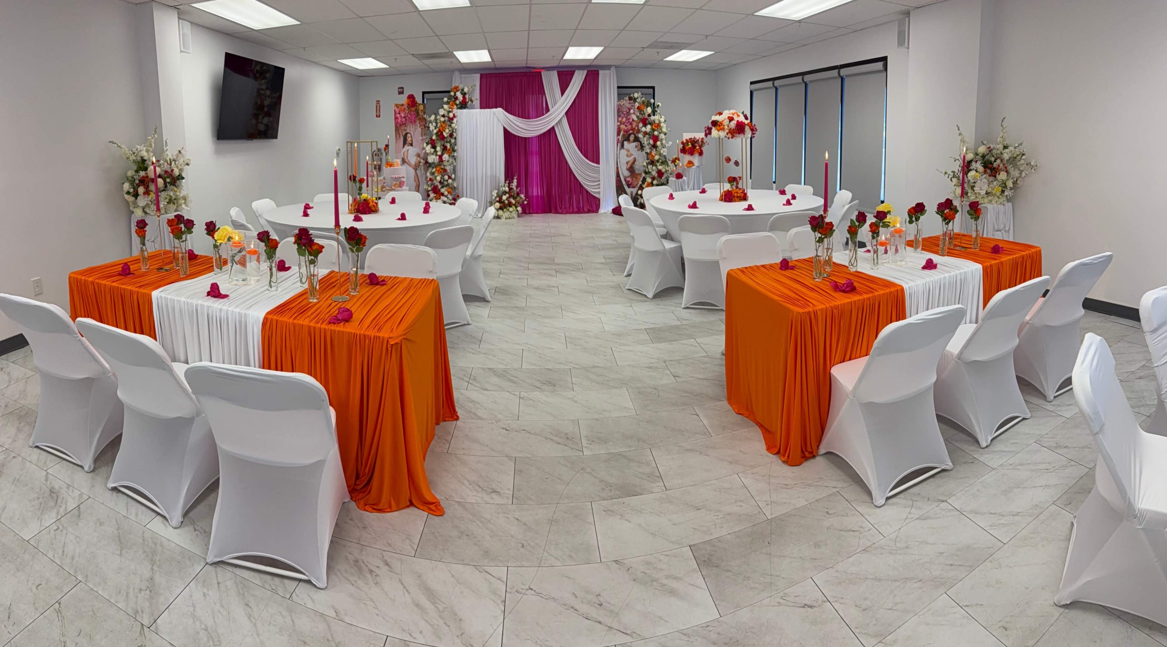 The image shows a decorated indoor event space with orange and white table linens, floral arrangements, and a backdrop featuring pink drapery.