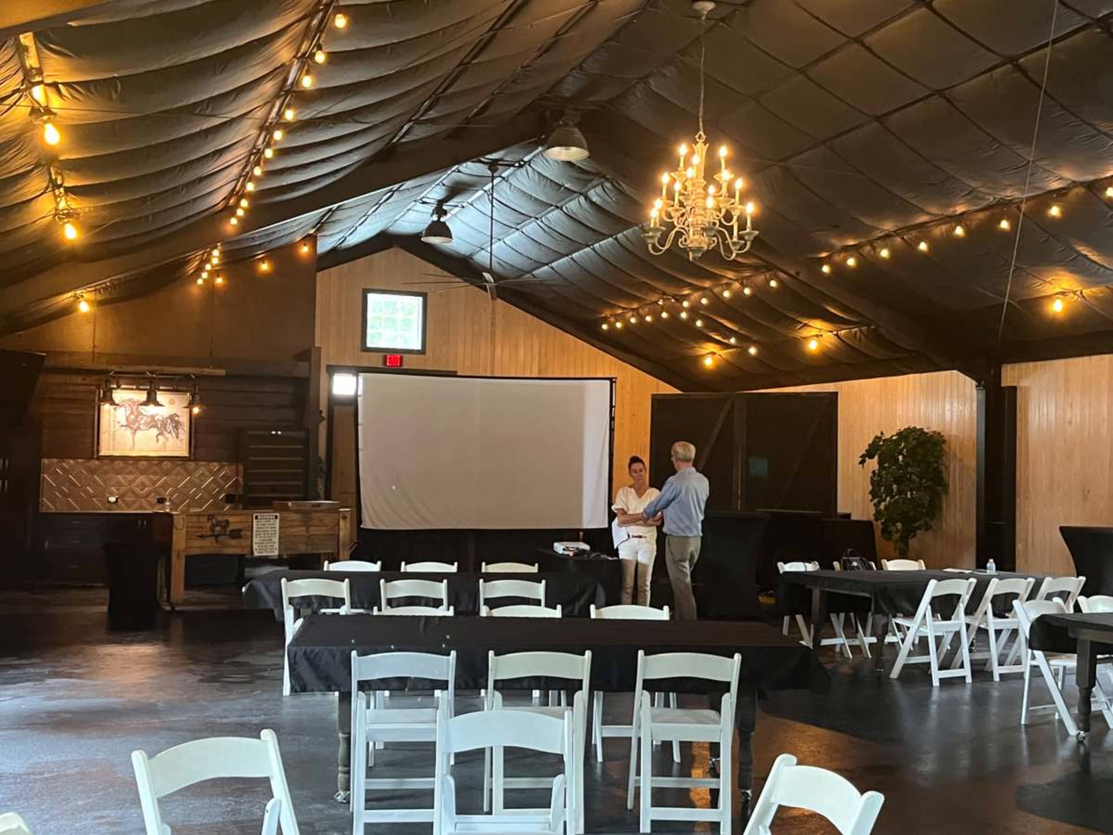 A spacious event venue features tables, chairs, and a large projection screen, with a couple discussing in the center under a chandelier.