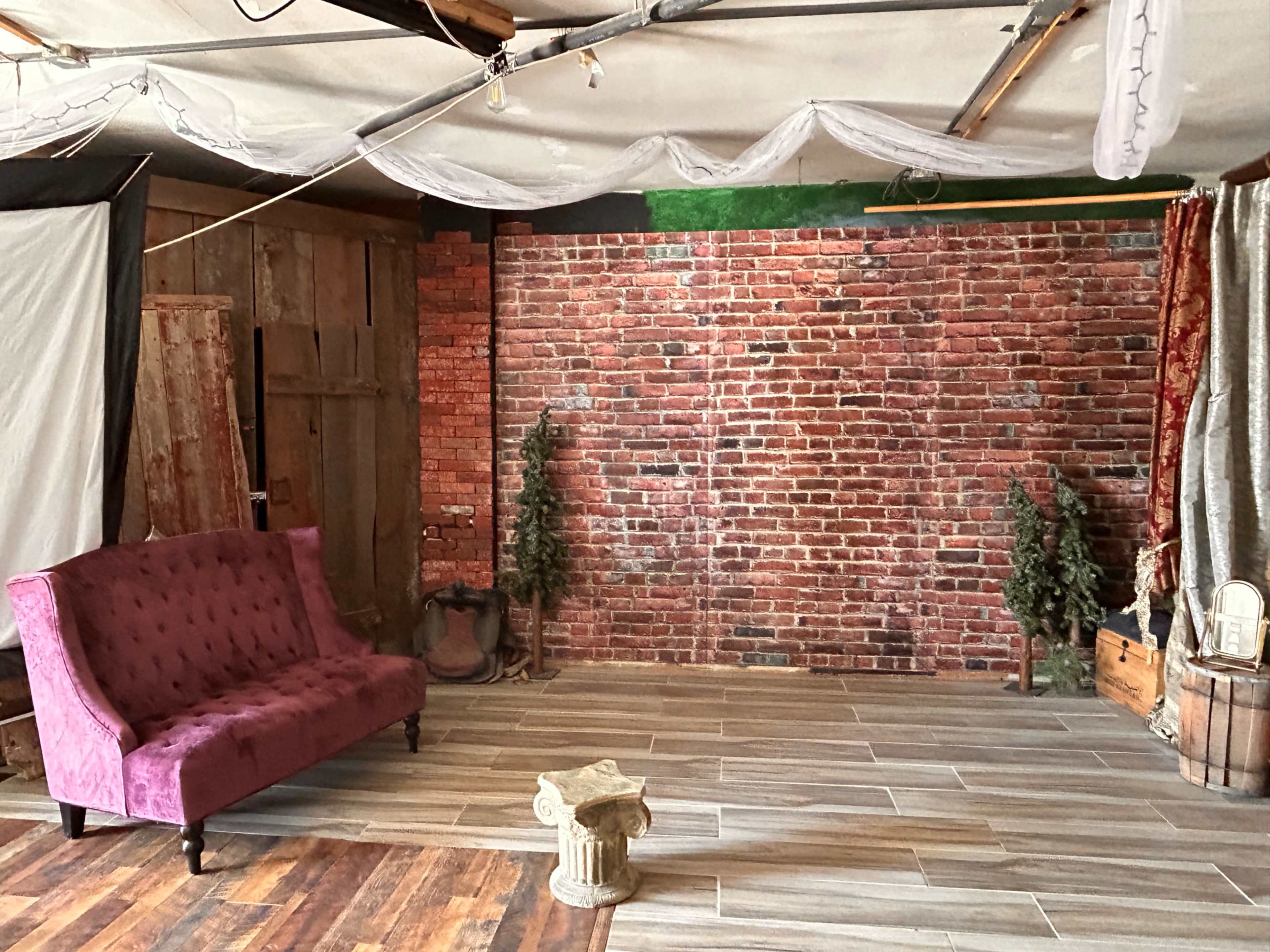 The image shows a studio space with a brick wall backdrop, a purple tufted sofa, and some decorative elements including potted trees and a small column.