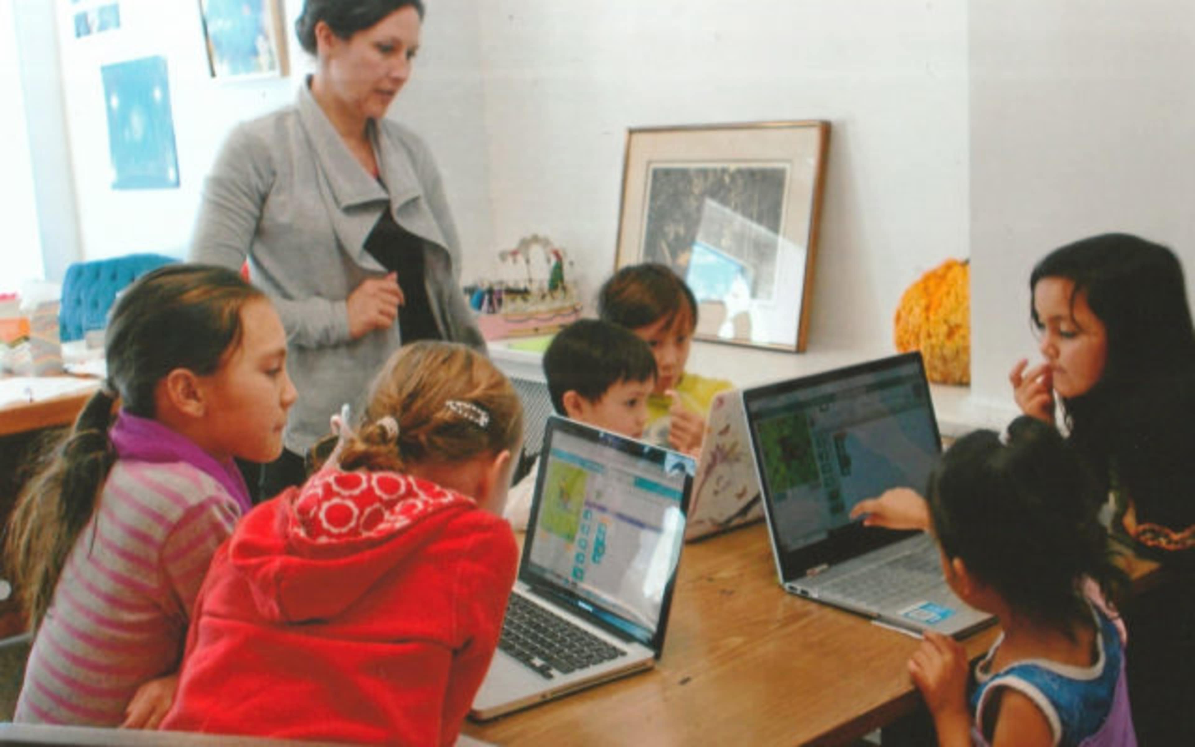 A group of children and an adult are engaged with laptops at a table in a classroom setting.