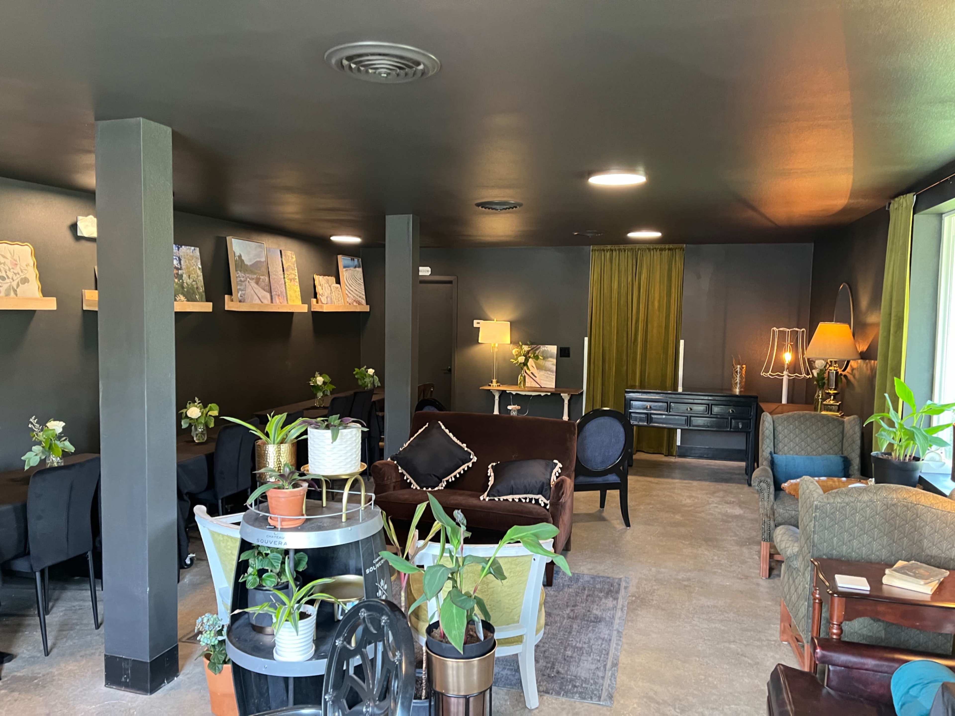 A cozy indoor space featuring various seating arrangements, plants, artwork on the walls, and warm lighting.
