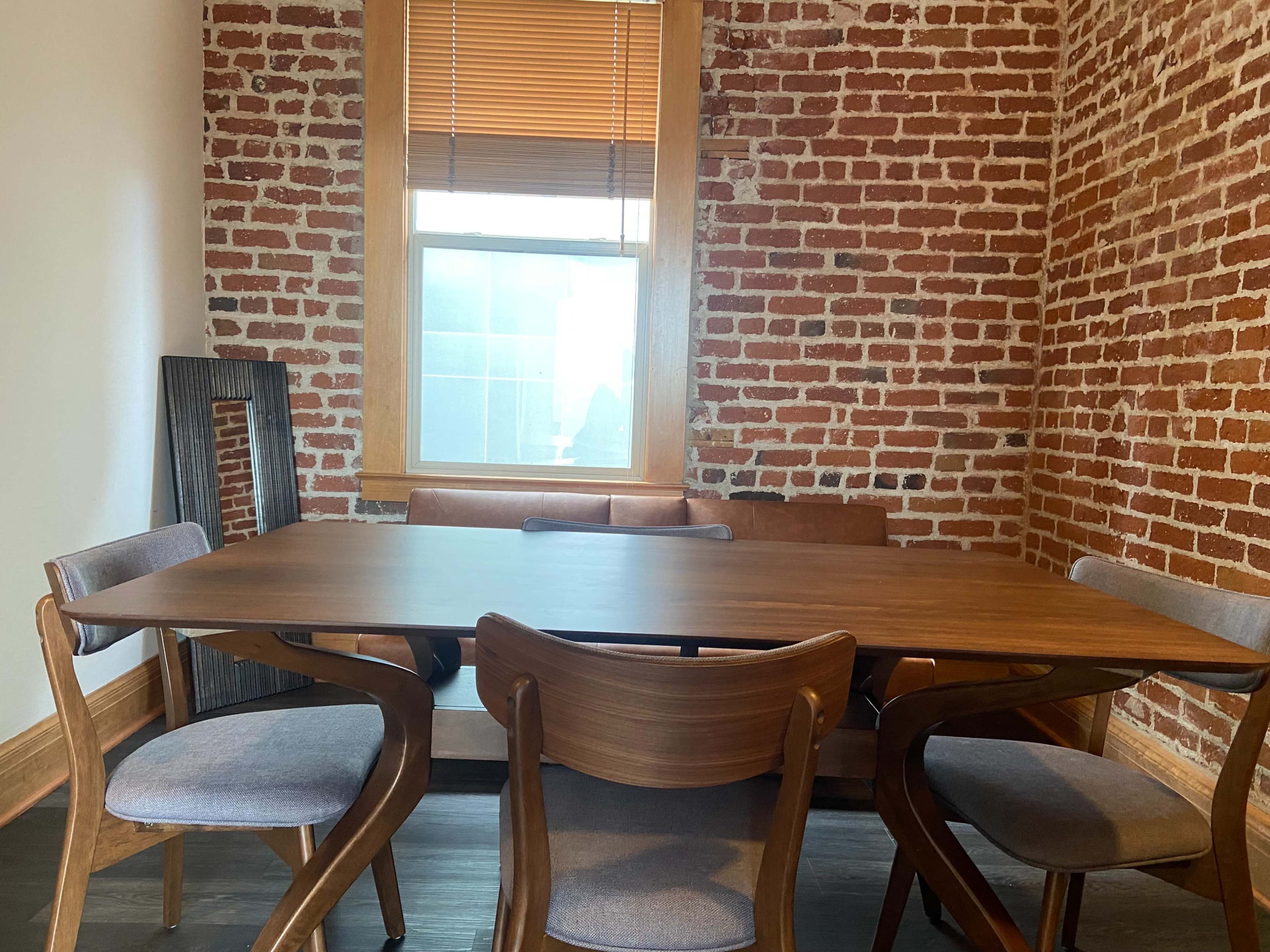 A wooden dining table with four chairs is positioned in a room featuring exposed brick walls and a large window.