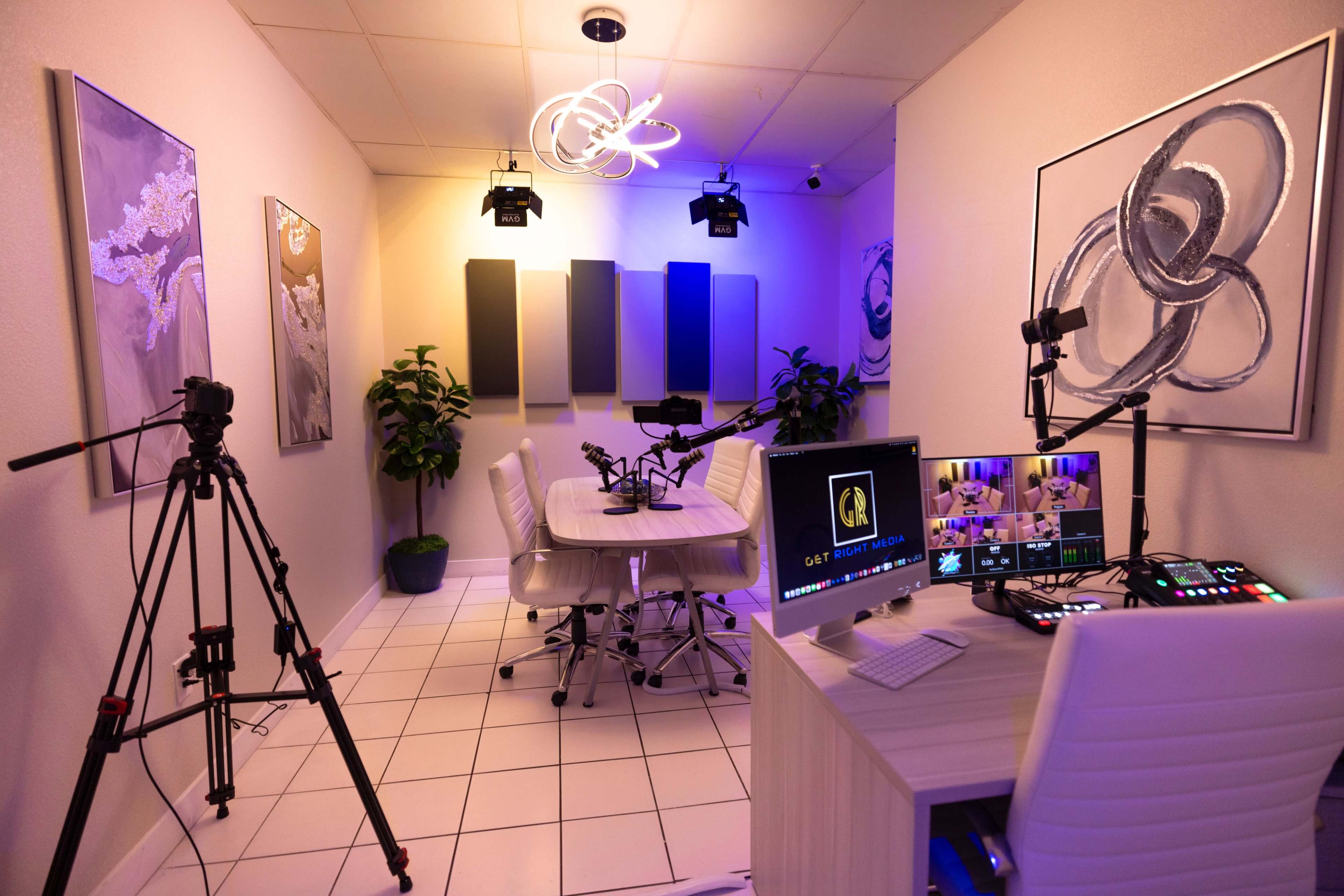 A recording studio with a round table, multiple cameras on tripods, and a computer setup displaying a live stream interface, illuminated by overhead lights.