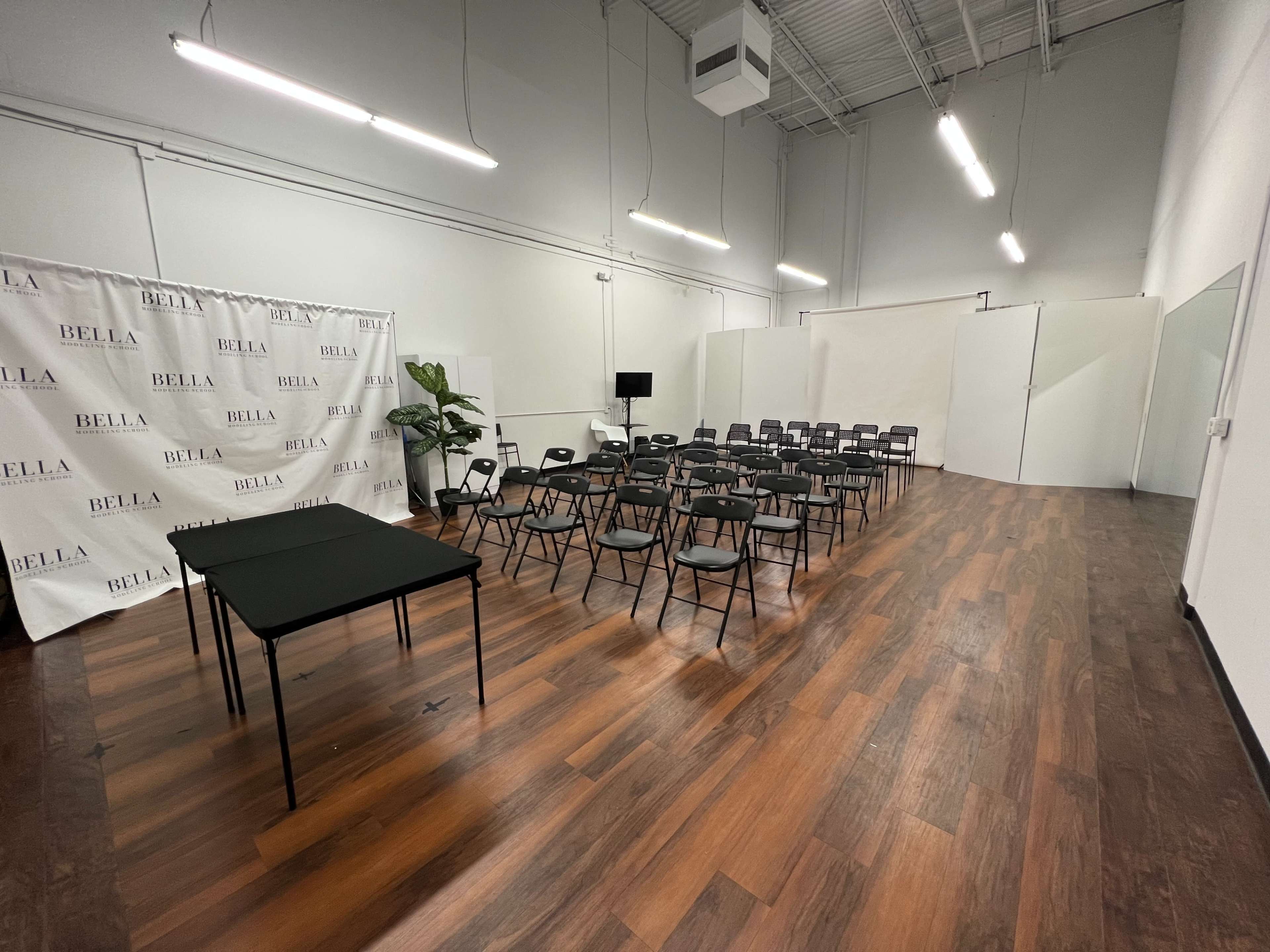 A spacious event room with a seating arrangement of black chairs facing a blank white backdrop, with two tables located at the front and a large mirror on one side.