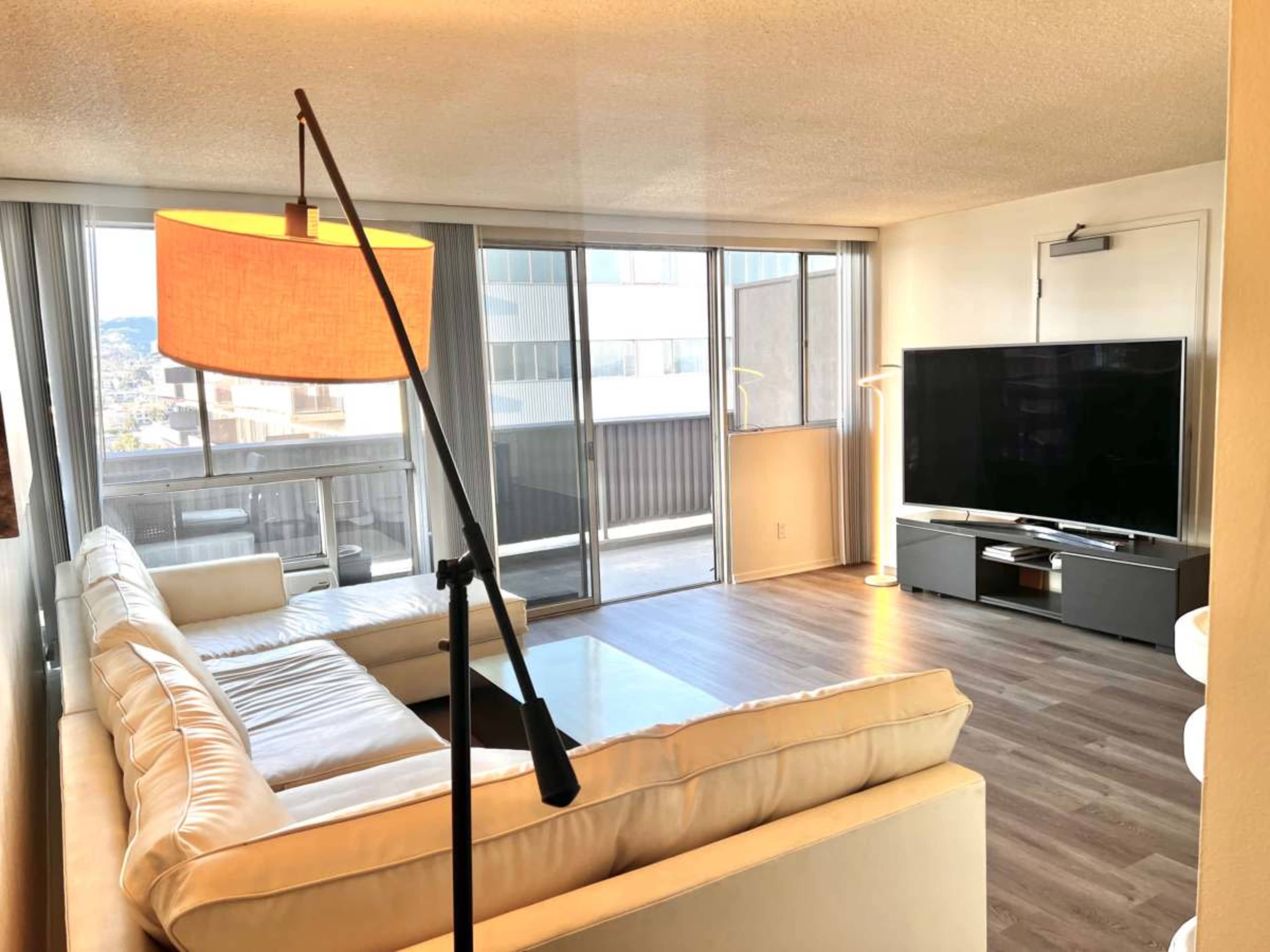 The image shows a modern living room with a large white sectional sofa, a floor lamp, and a flat-screen television against a wall of windows letting in natural light.