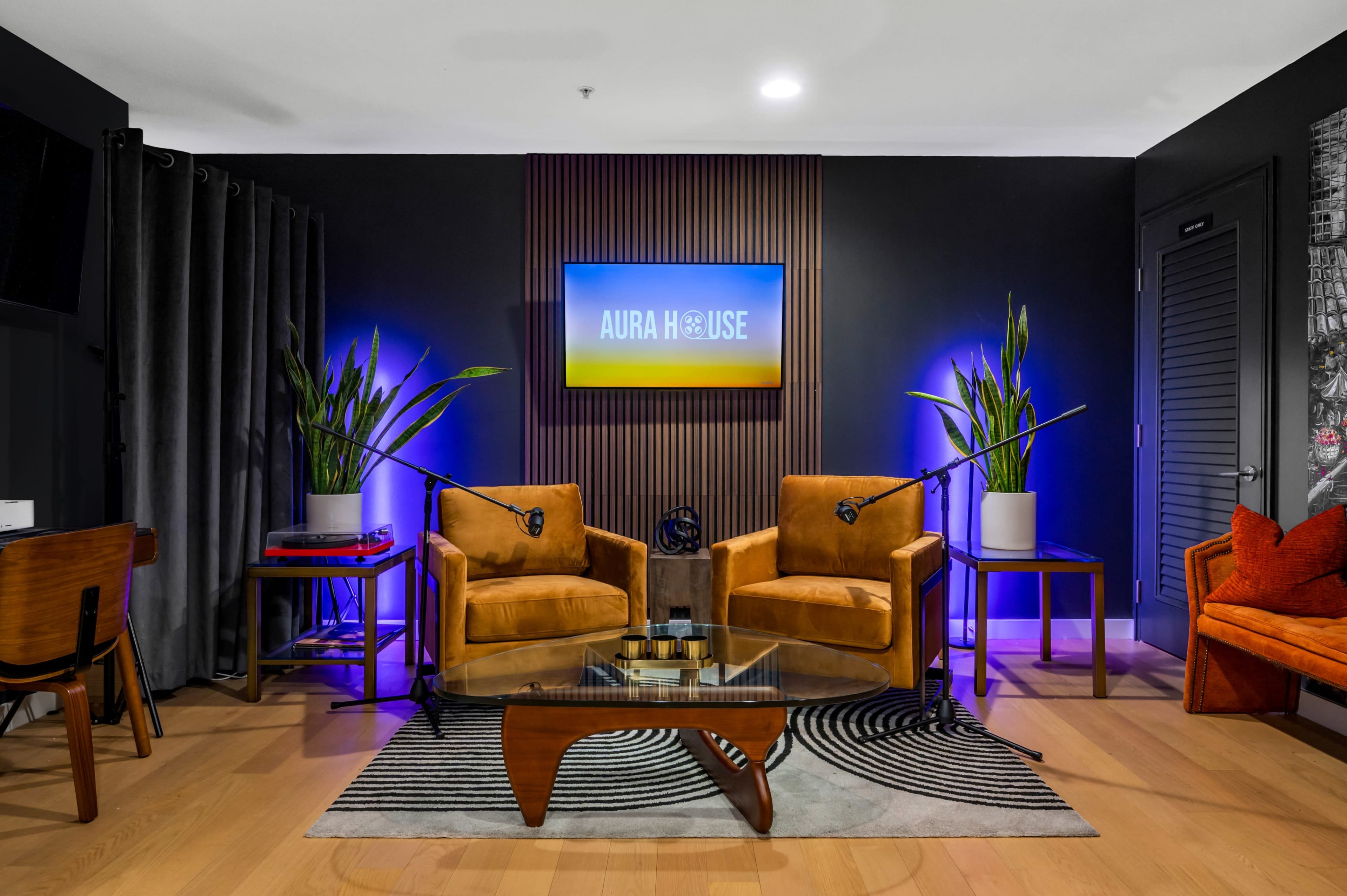 A modern lounge area with two brown chairs, a glass coffee table, and a flat-screen TV mounted on the wall, accompanied by potted plants and colorful lighting.