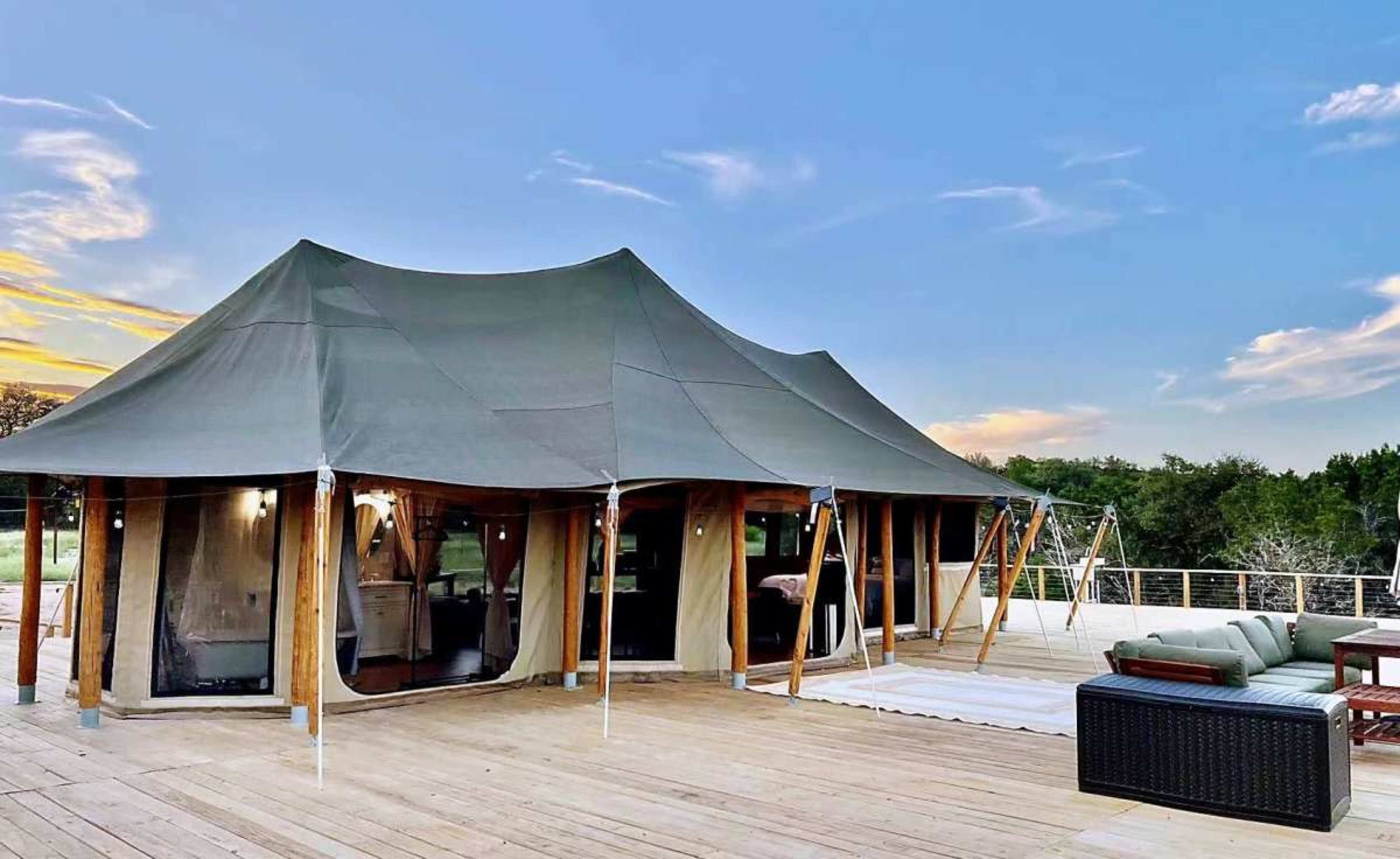 A large canvas tent with a wooden deck is set against a clear sky and green trees.