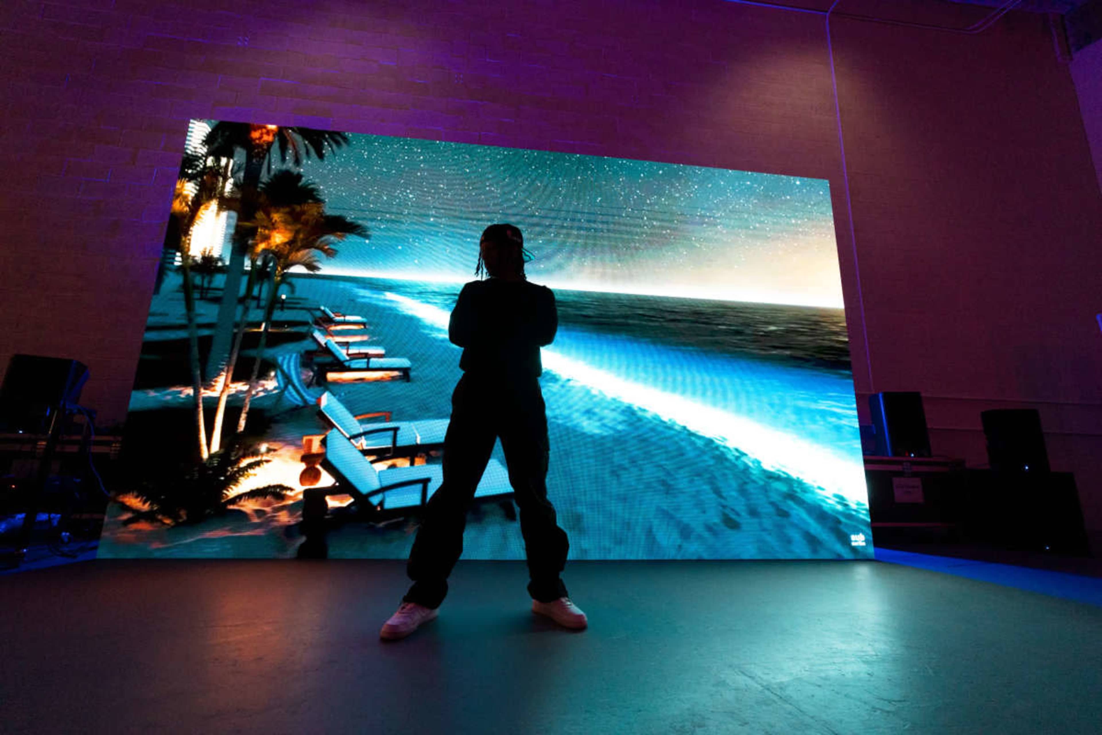 A person stands in front of a large screen displaying a nighttime beach scene with lounge chairs and ocean waves.