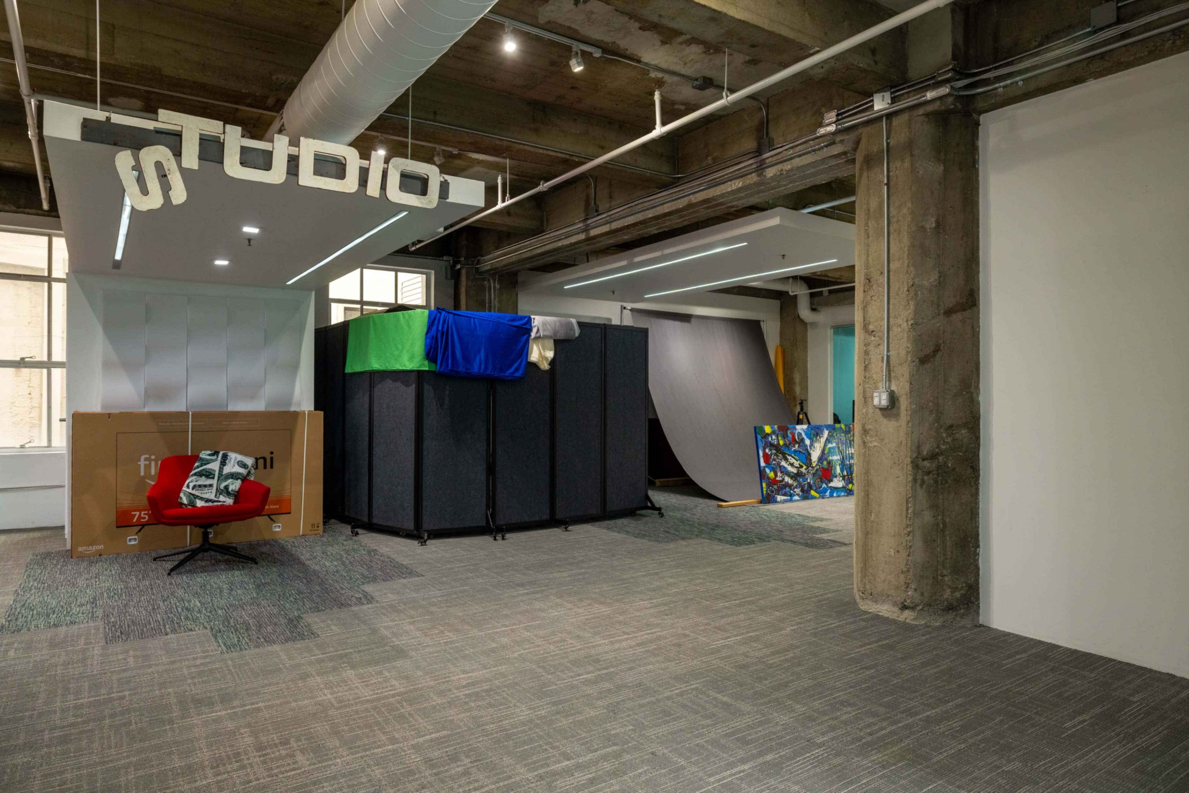 The image shows an industrial-style interior space labeled "STUDIO," featuring a combination of cardboard boxes, fabric draped over furniture, and large panels leaning against the walls.