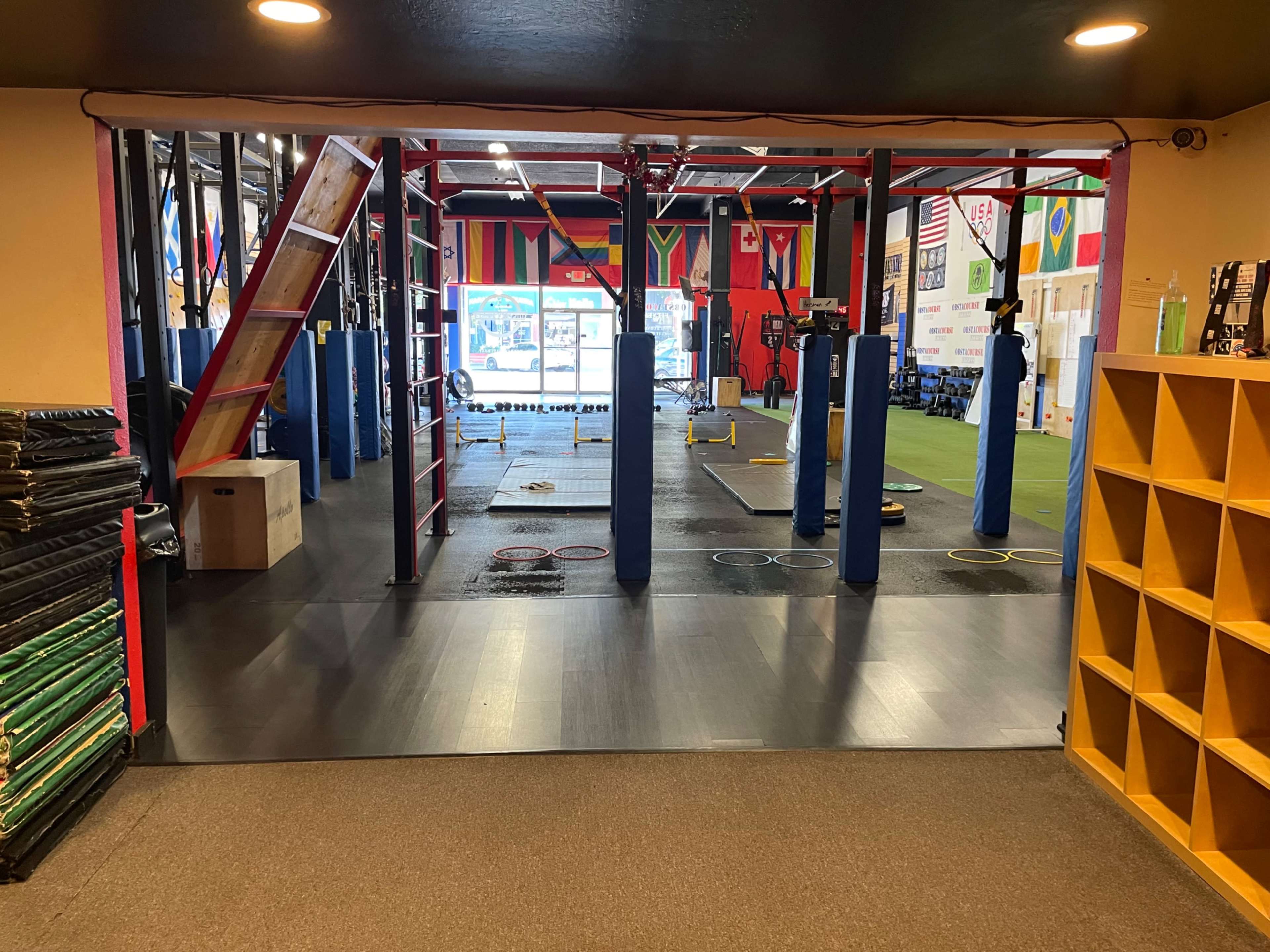 The image shows an indoor gym with various fitness equipment, including suspended training apparatus and colored rubber mats, surrounded by flags on the walls.