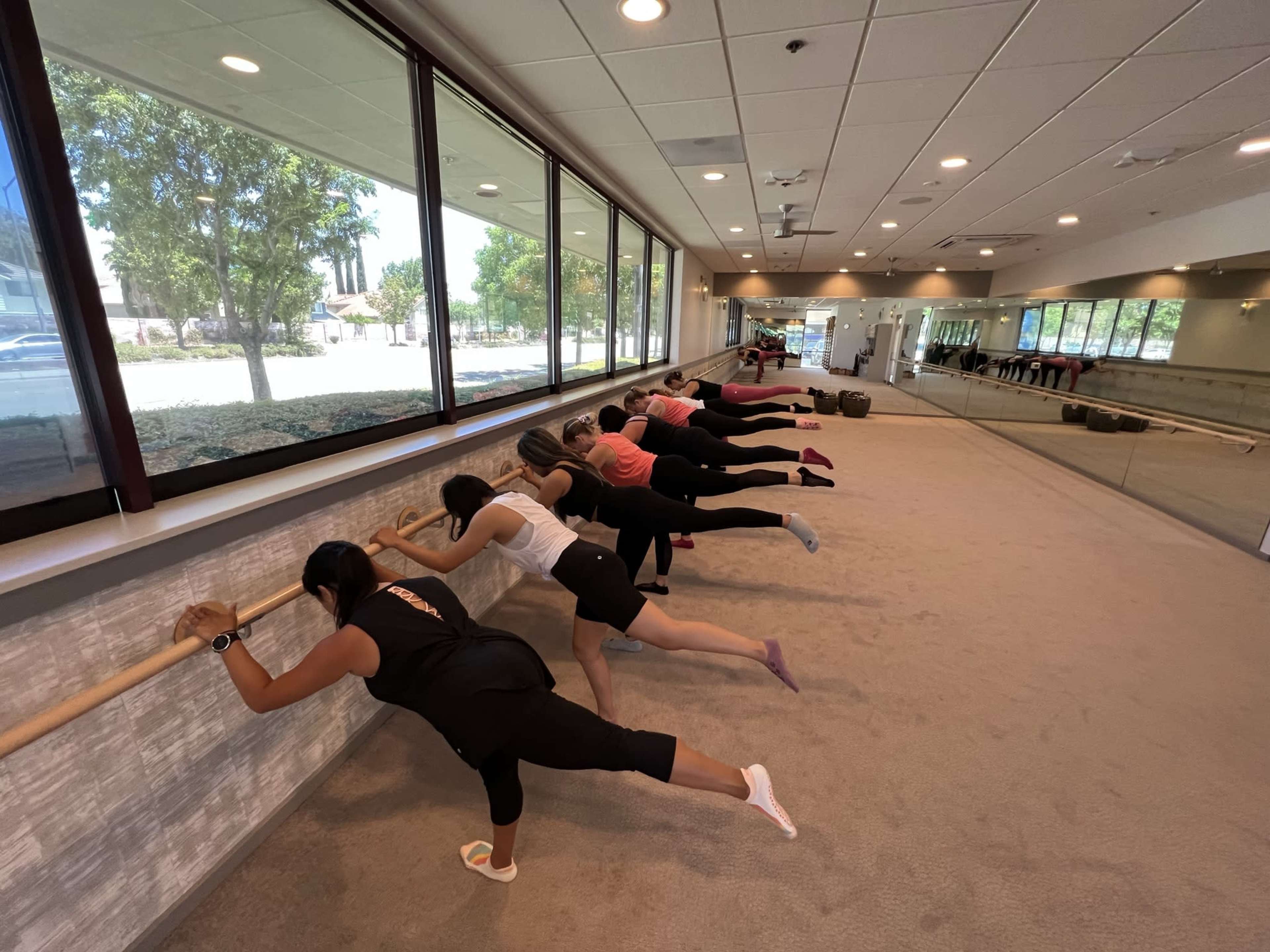 A group of individuals is performing barre exercises along a wooden ballet bar in a spacious studio with large windows.