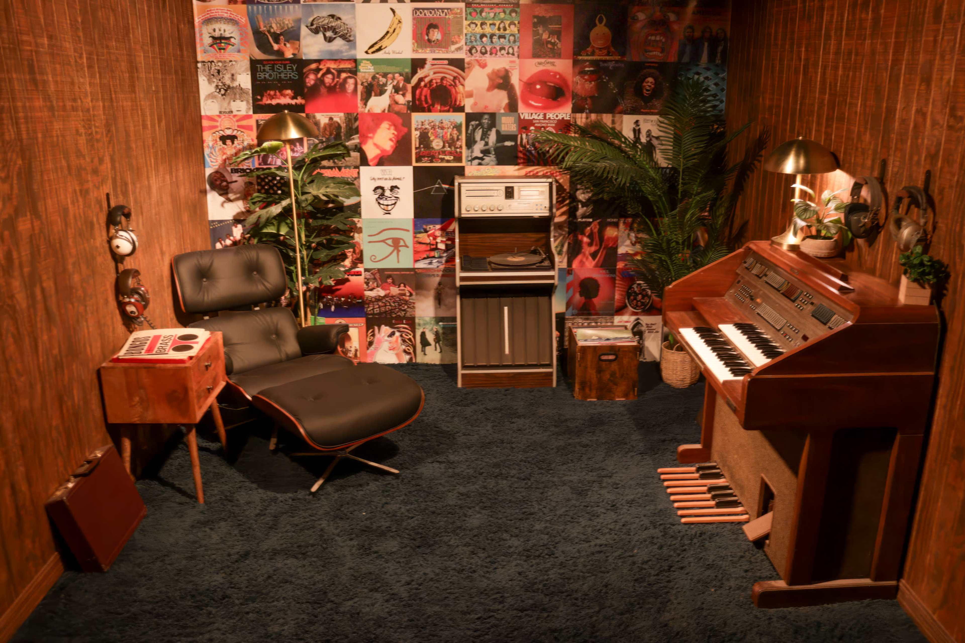 The image shows a retro-themed room with a vinyl record wall, a chair, a small table, a record player, and an organ surrounded by decorative plants.