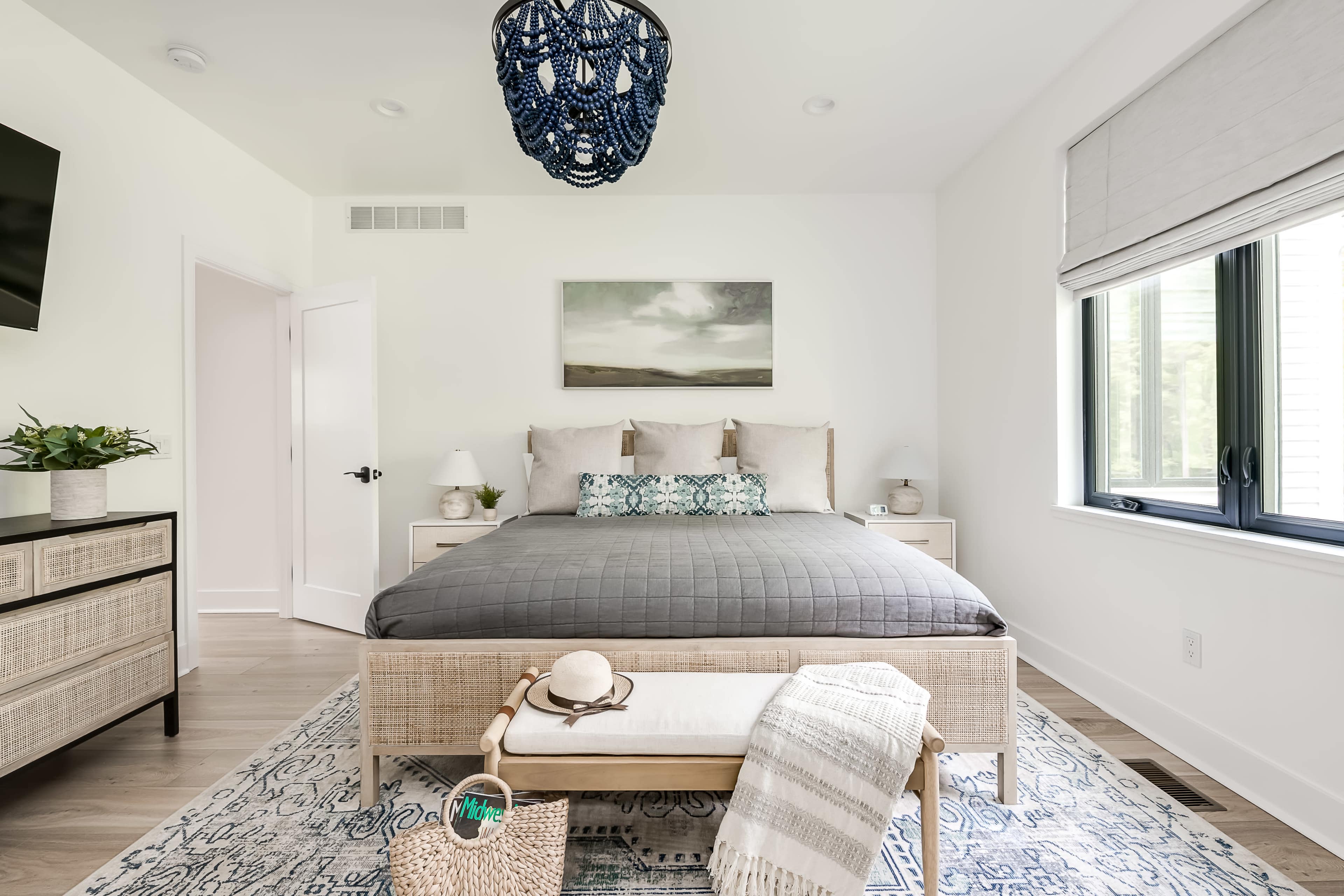 A modern bedroom features a large bed with gray bedding, two nightstands, a decorative rug, and a woven storage basket at the foot of the bed, illuminated by a blue pendant light.