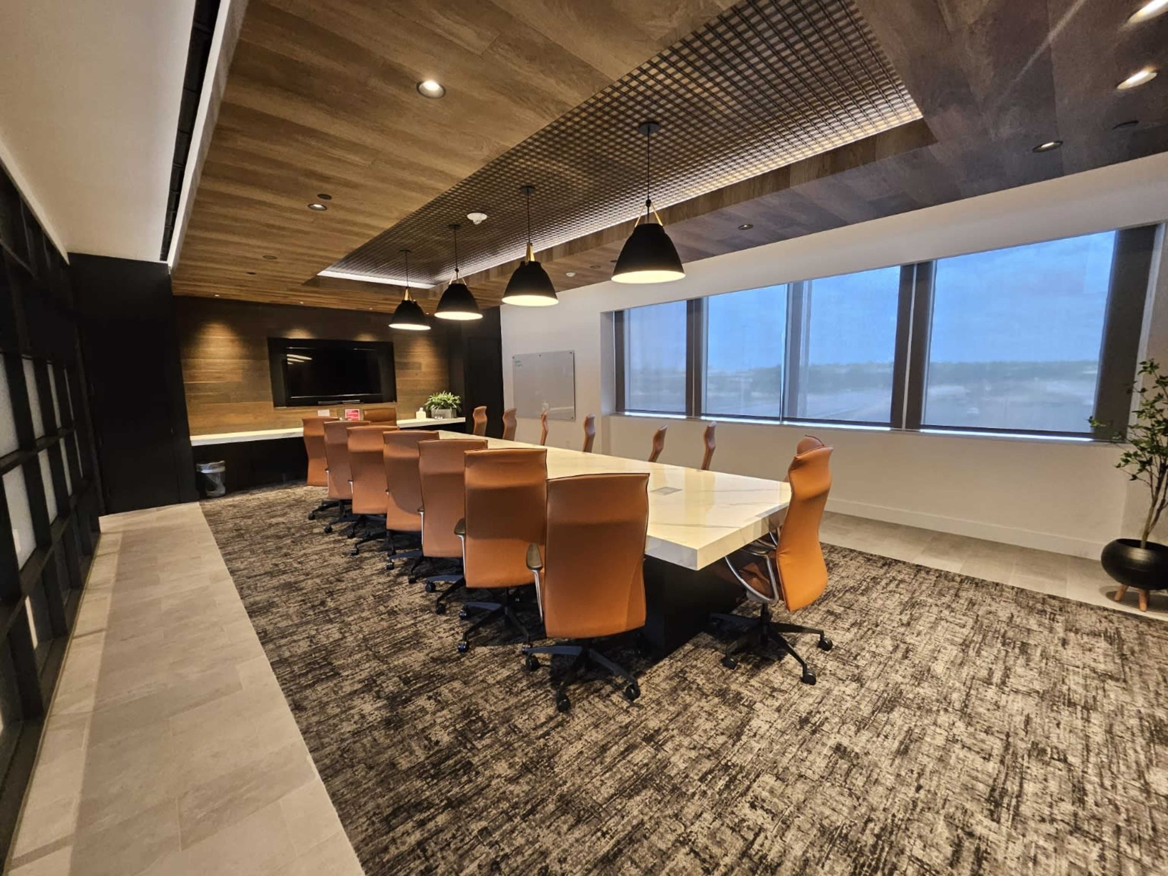 The image shows a modern conference room with a long white table, orange chairs, wooden ceiling panels, and large windows.