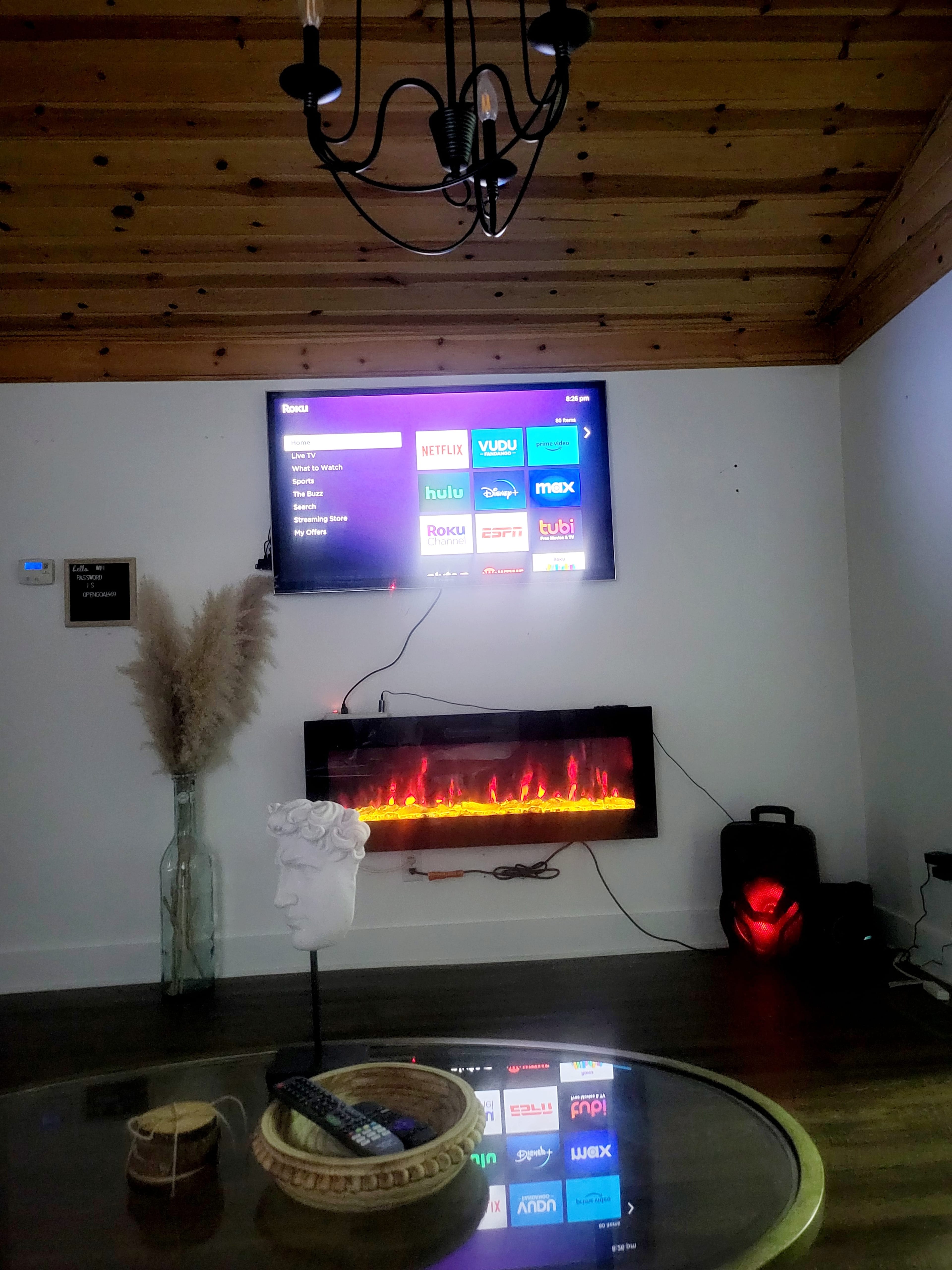 A living room features a mounted television displaying streaming app options and an electric fireplace below, with a decorative vase and remote on a round coffee table.