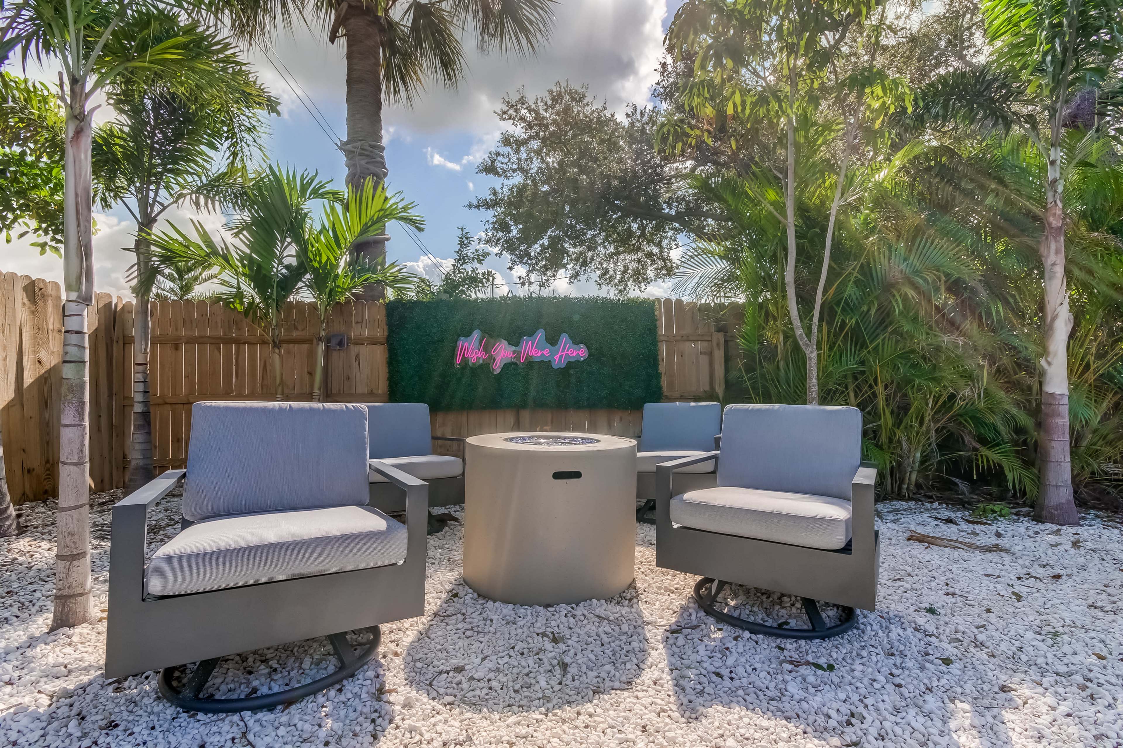 A seating area featuring four gray chairs arranged around a circular table, surrounded by palm trees and a green wall sign that says "Make You Feel At Home."