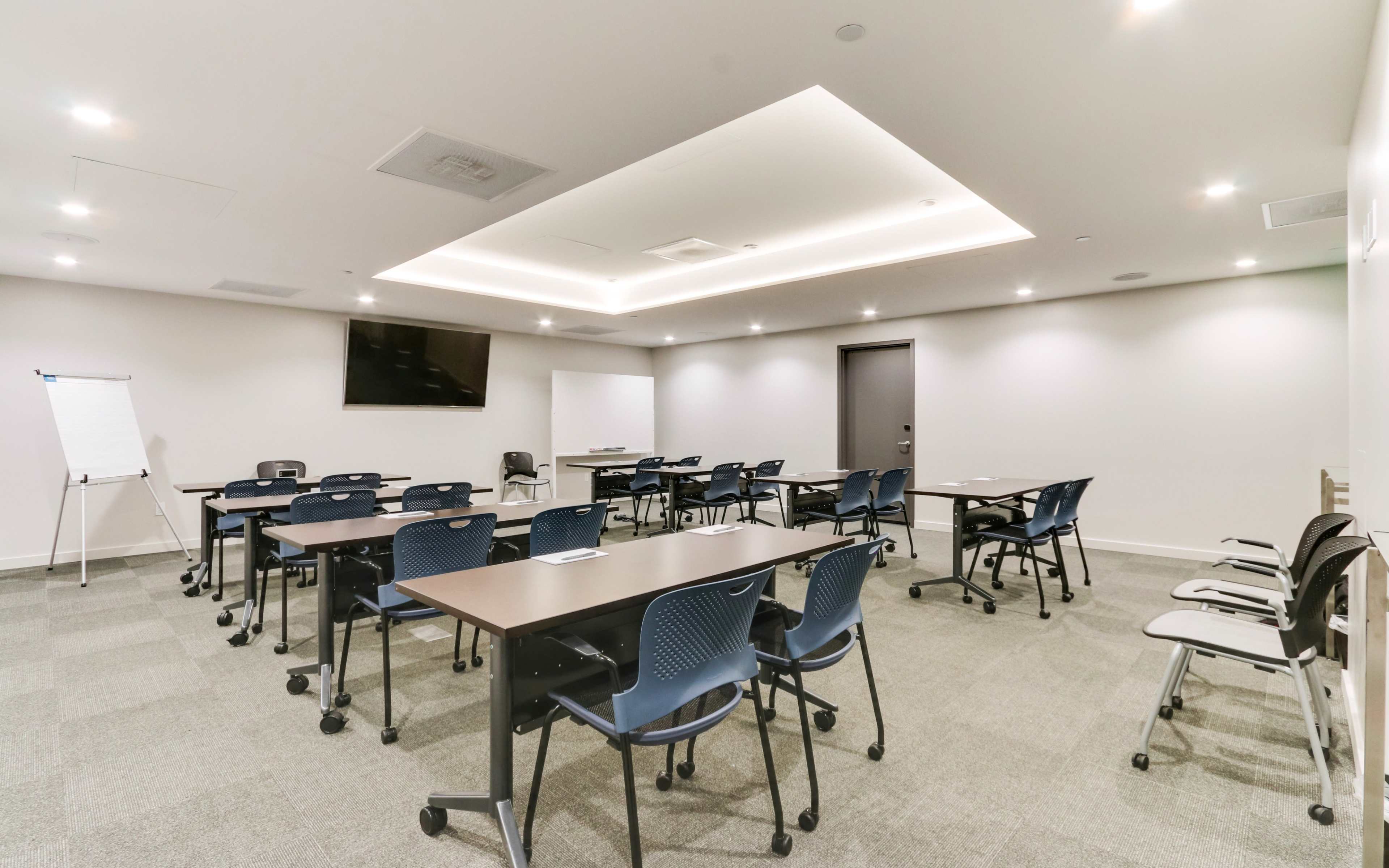 A modern conference room is arranged with several tables and chairs, featuring a flat-screen TV and a whiteboard.