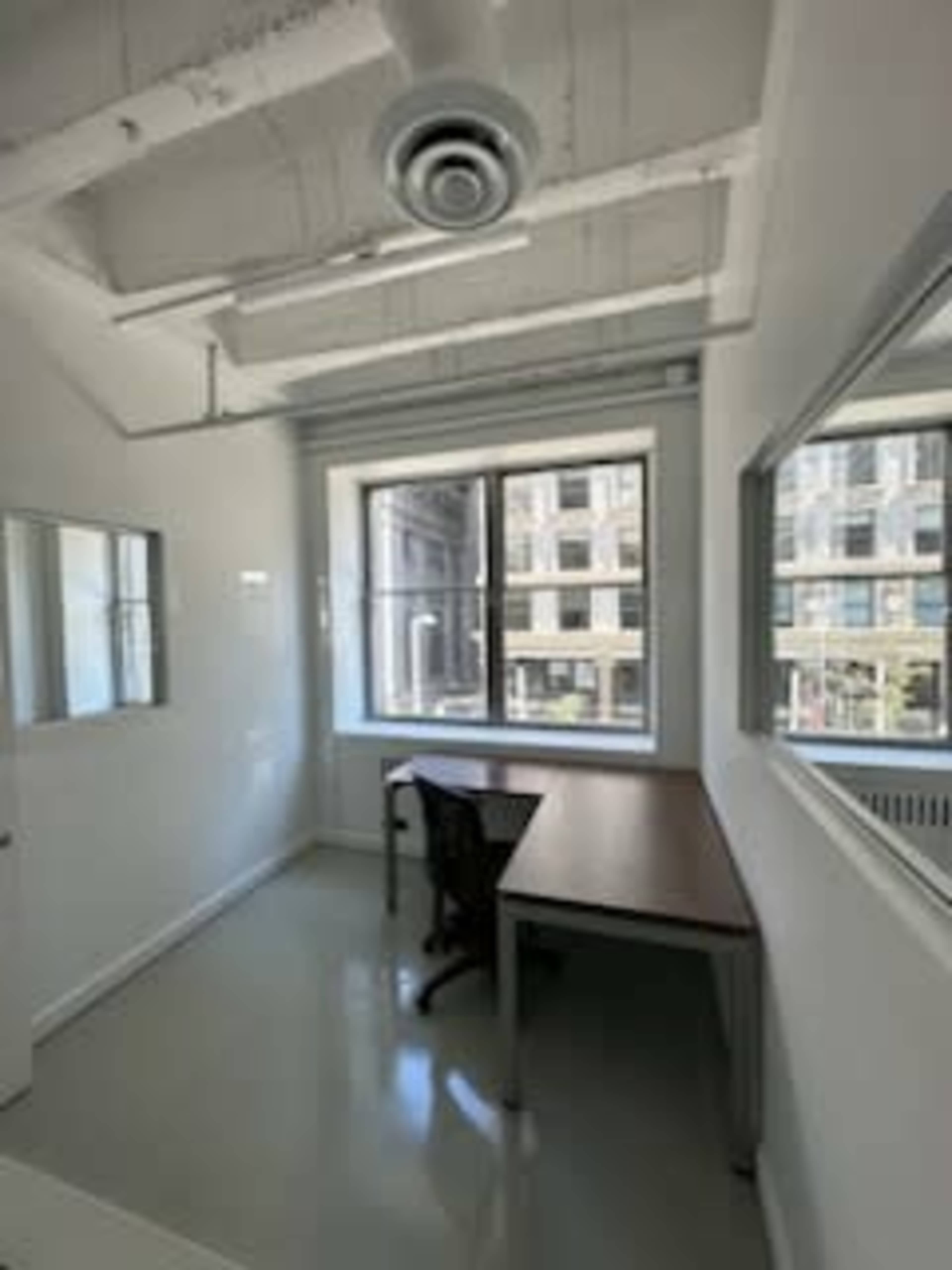 The image shows a small, empty office space with a desk and a chair, white walls, and a large window overlooking a city view.