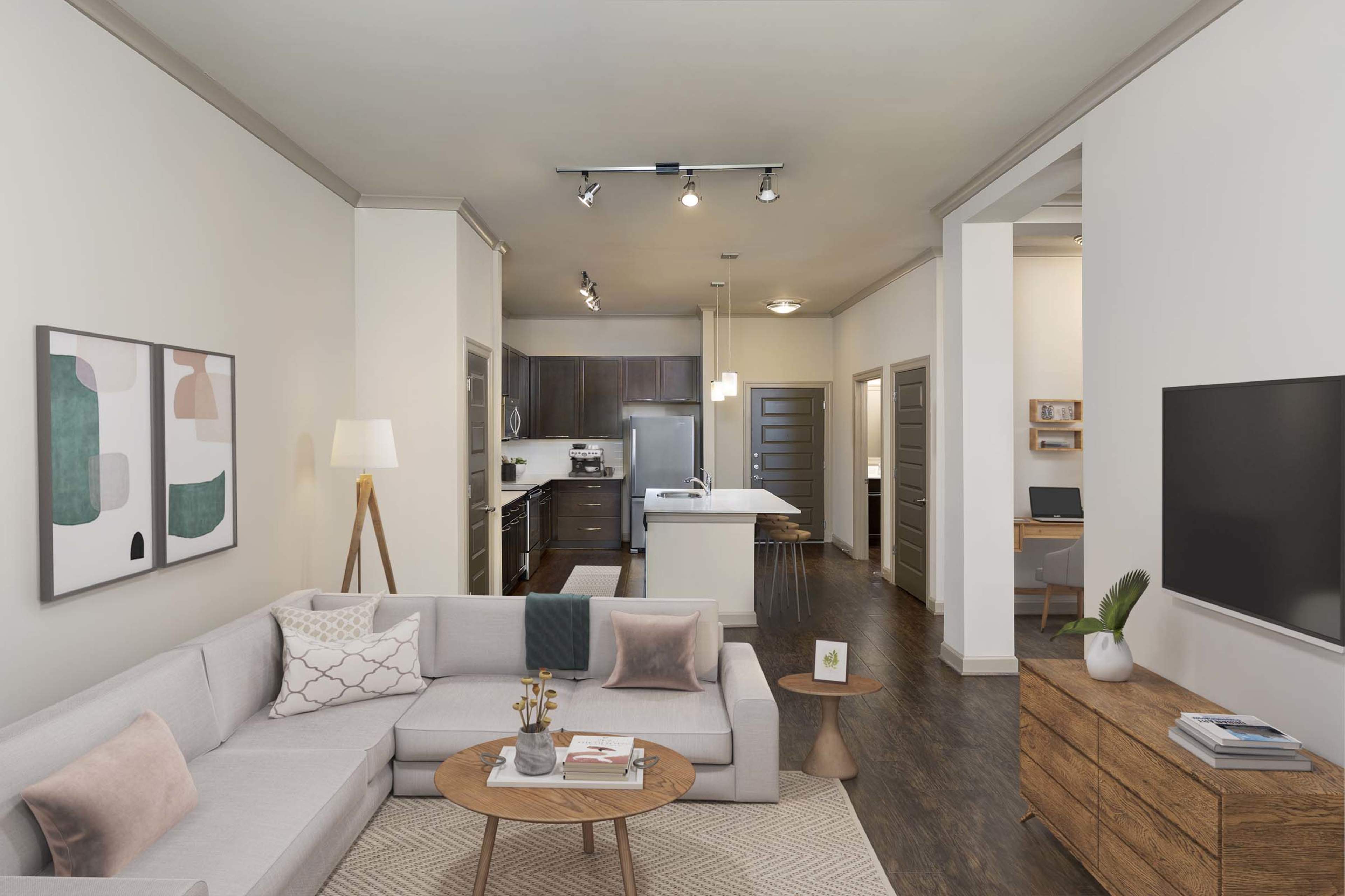 The image shows a modern living room featuring a light gray sectional sofa, a round coffee table, and a television mounted on the wall, with a kitchen visible in the background.