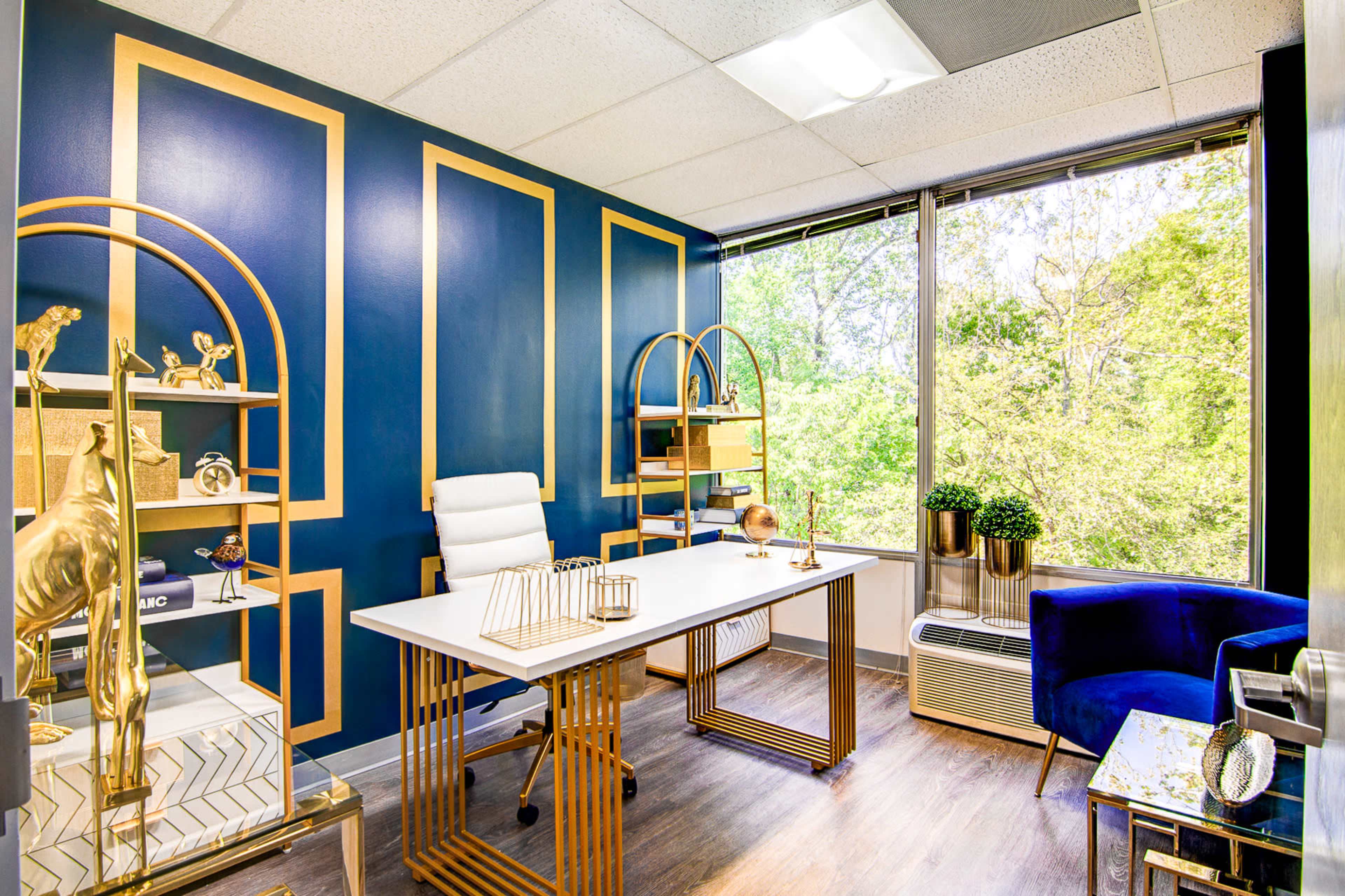 A modern office space features a desk with a white chair, gold accents, and large windows overlooking greenery.