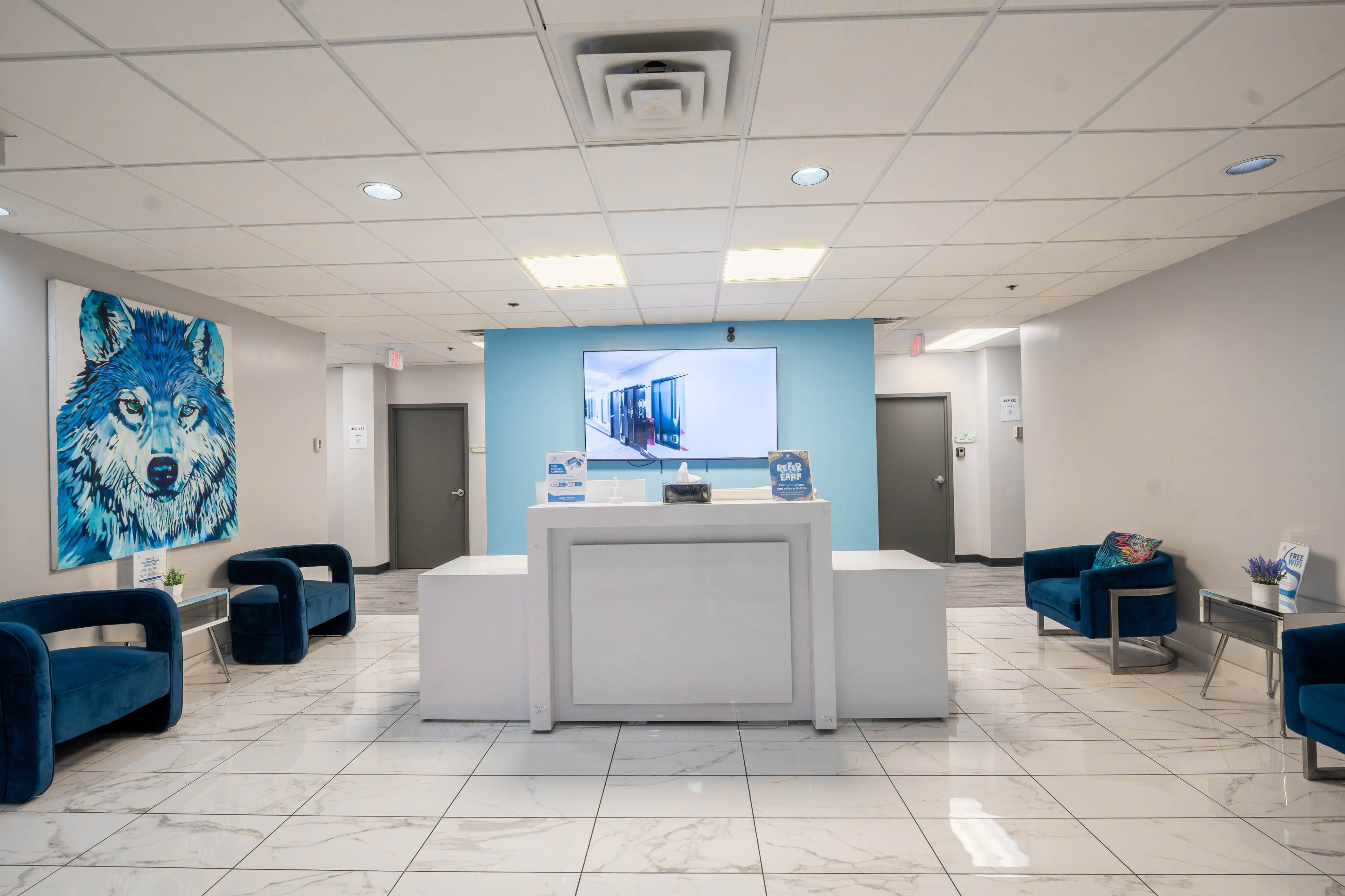 The image shows a modern waiting area with a reception desk, blue accent walls, and plush seating in a well-lit environment.