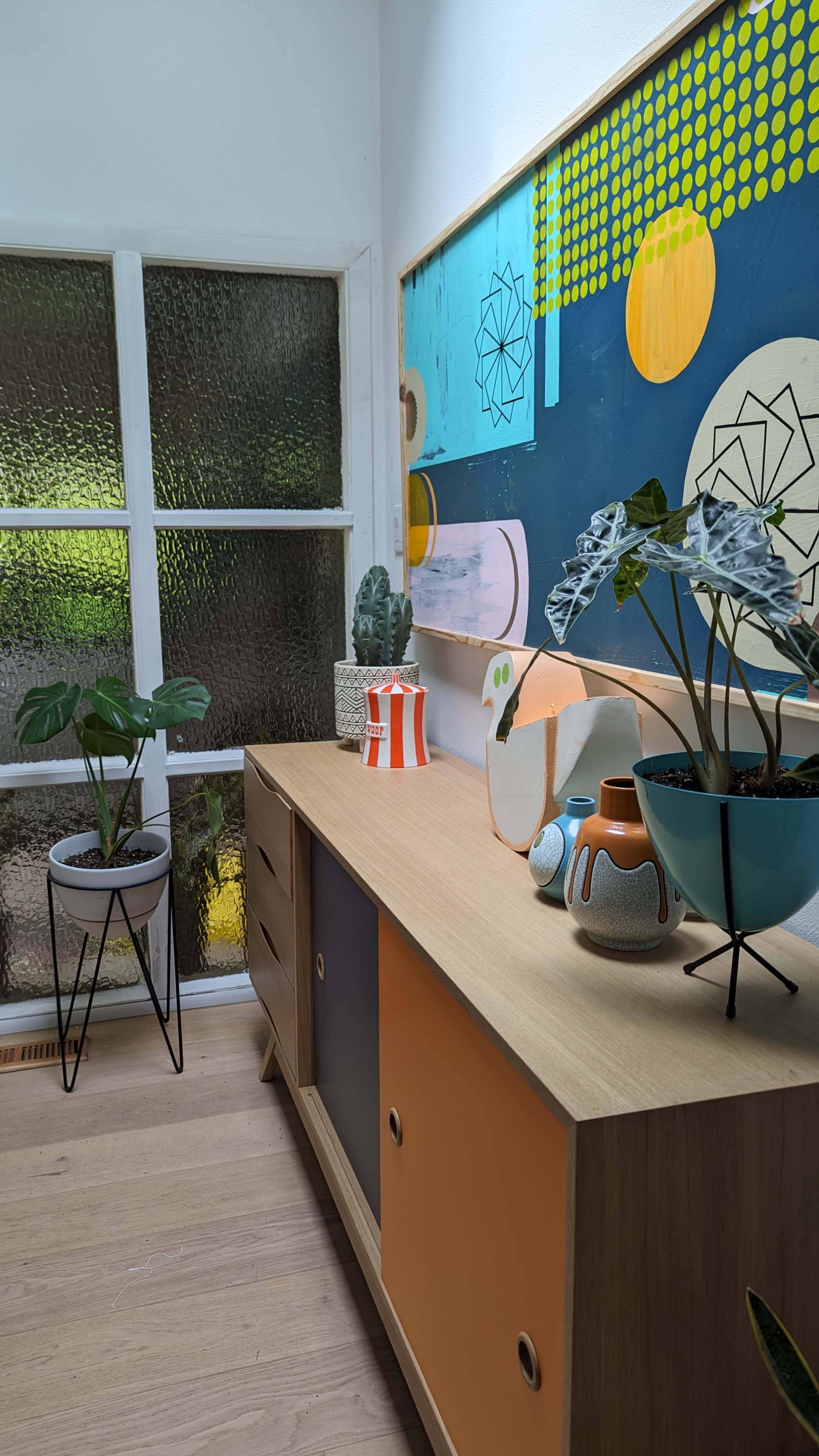 The image shows a modern wooden sideboard adorned with decorative plants and colorful ceramic vases, positioned near a textured glass window.