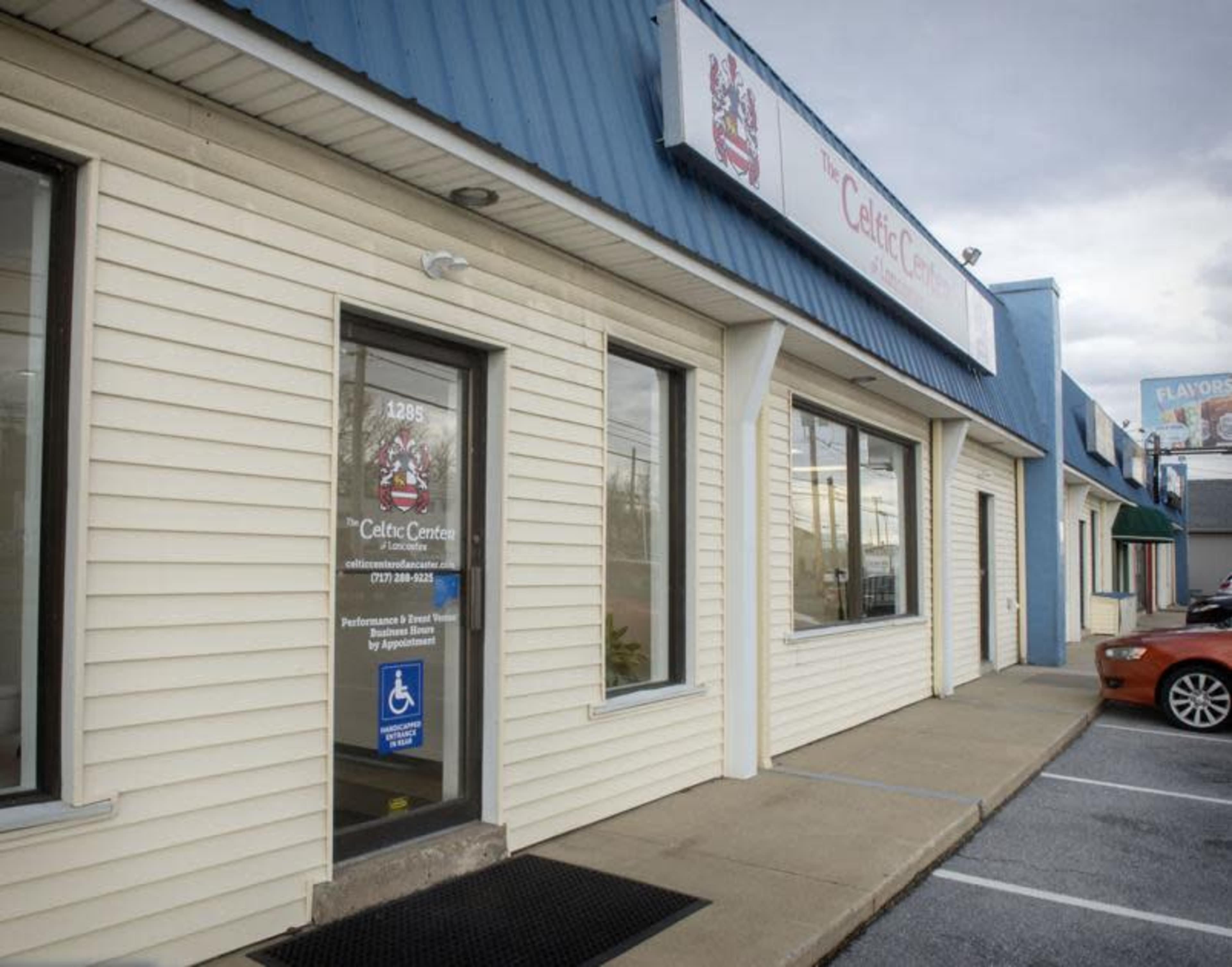 A single-story commercial building with a light-colored exterior and a blue metal roof, featuring multiple storefronts and a parking area.