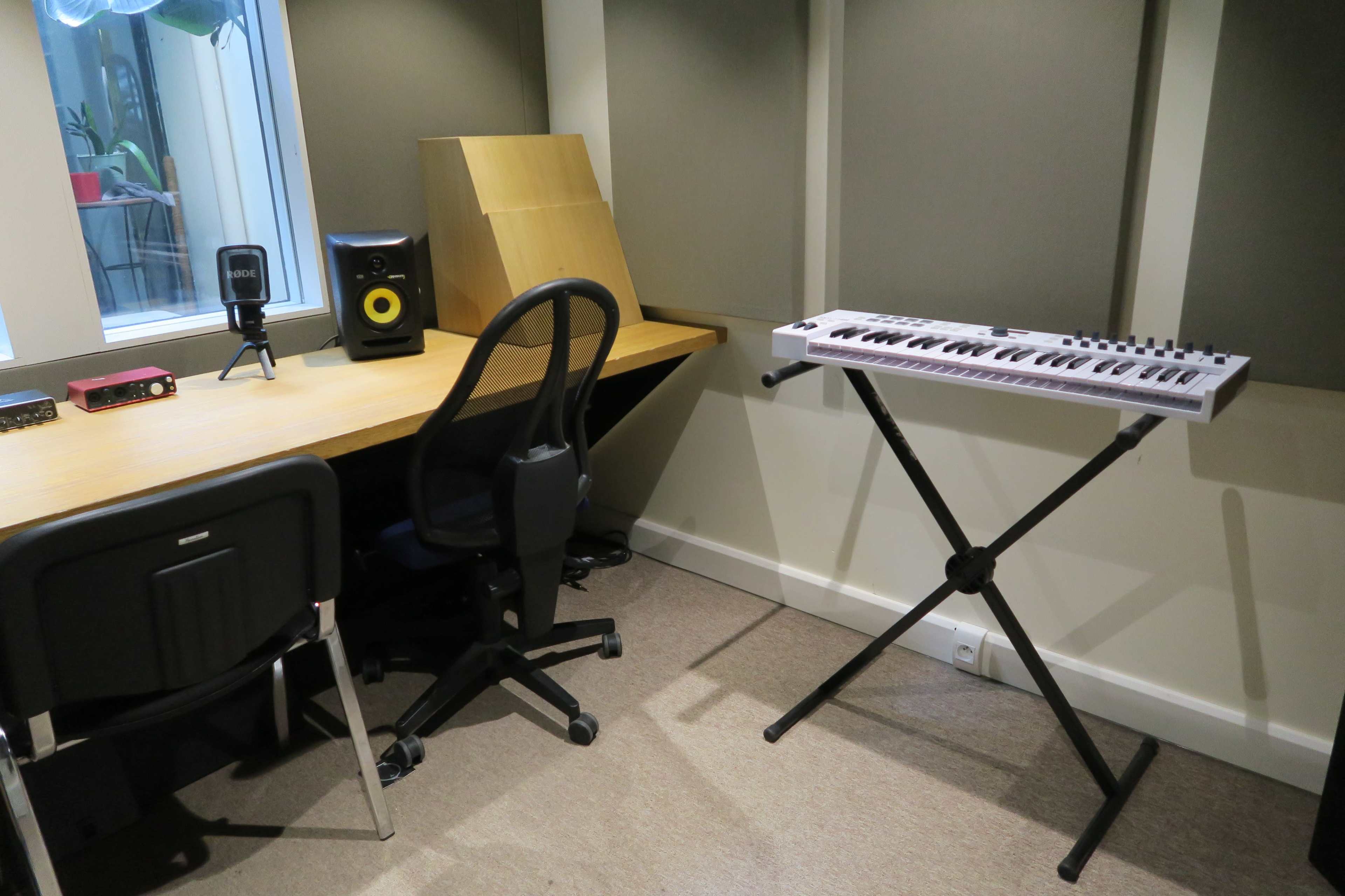 The image shows a small music production room with a desk, a chair, studio monitor speakers, and a keyboard on a stand.
