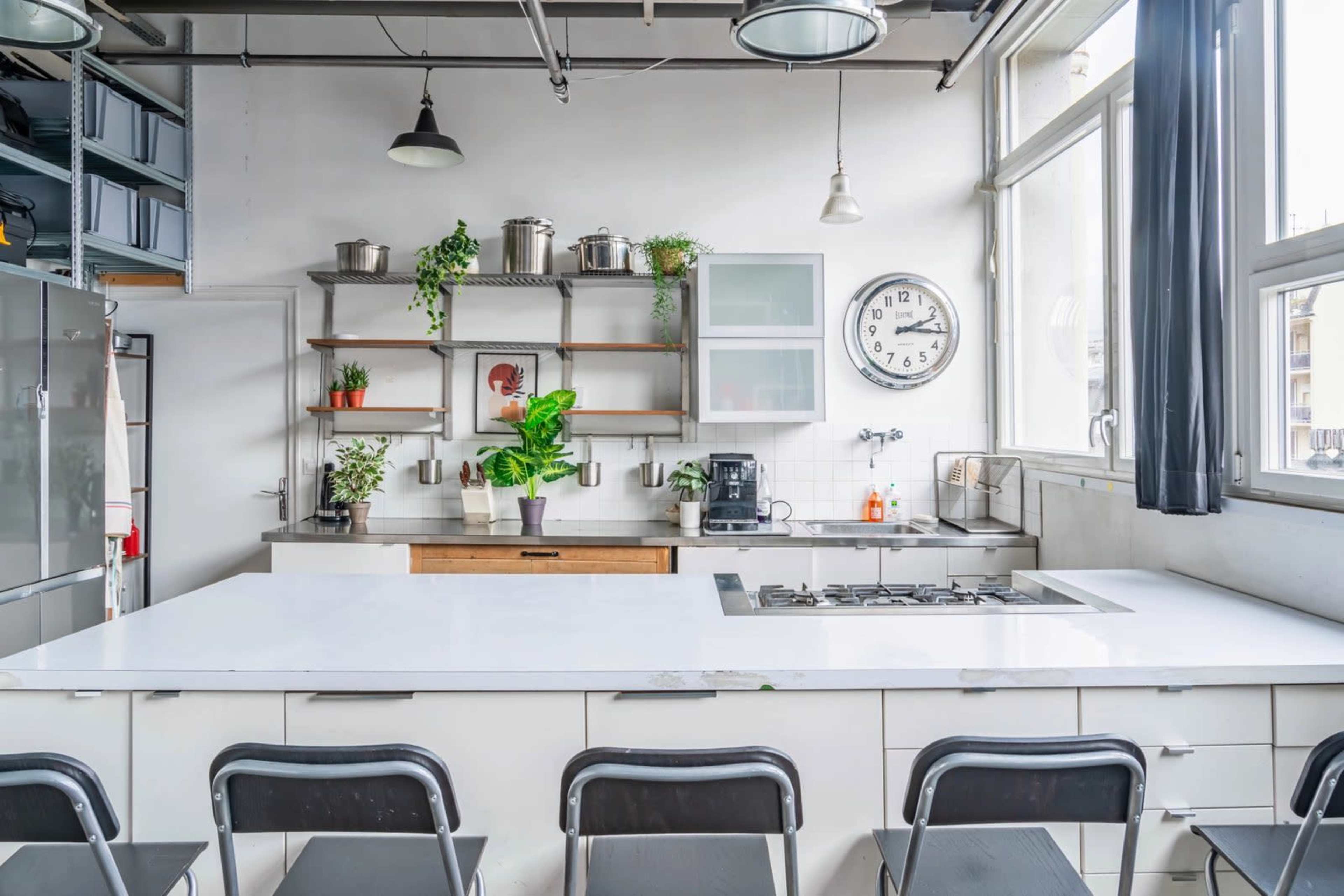 The image shows a modern kitchen with a white countertop, open shelving with plants and kitchenware, and large windows allowing natural light.