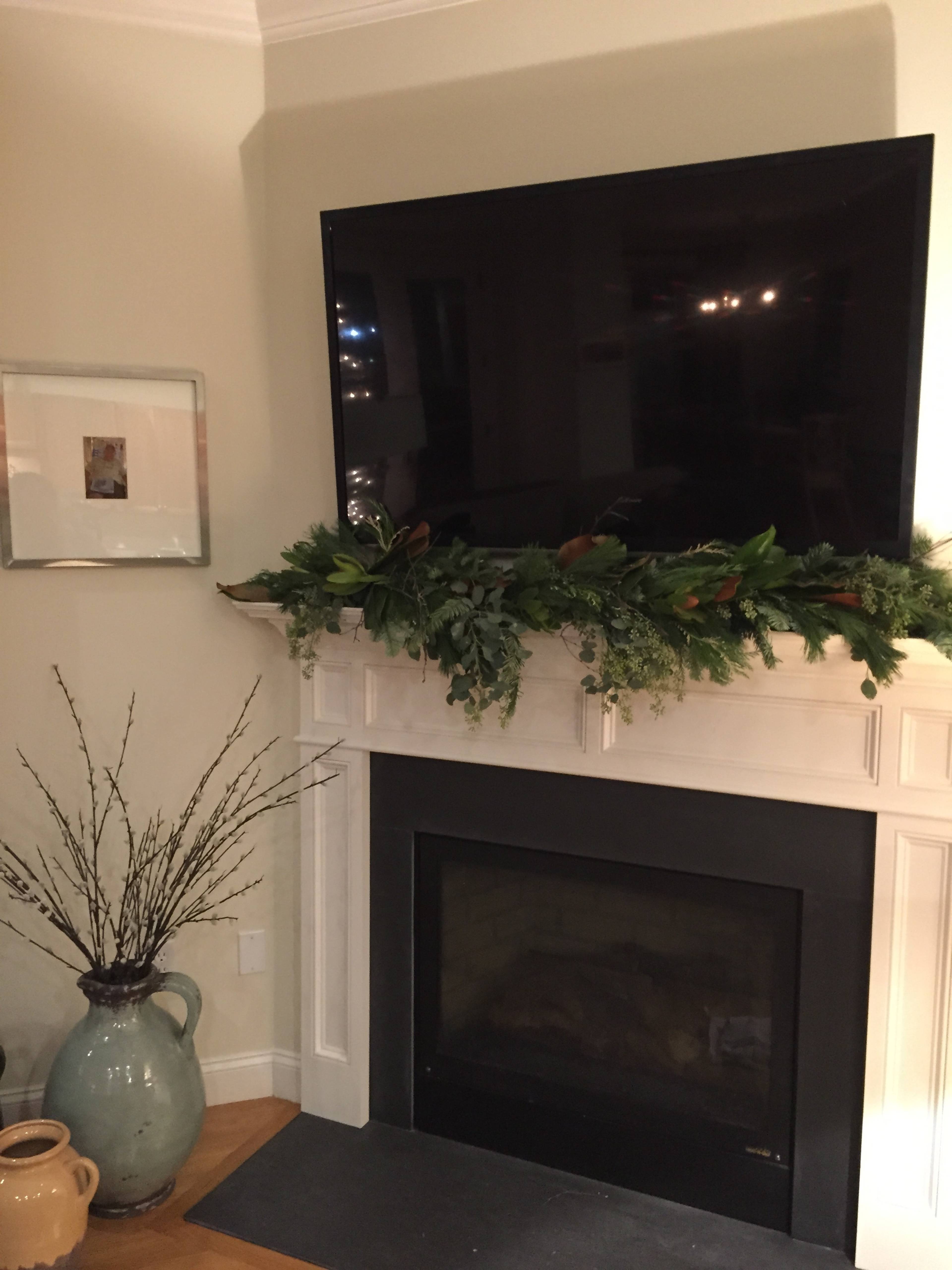 A flat-screen television is mounted above a mantel adorned with greenery, above a black fireplace with a simple vase and branches nearby.