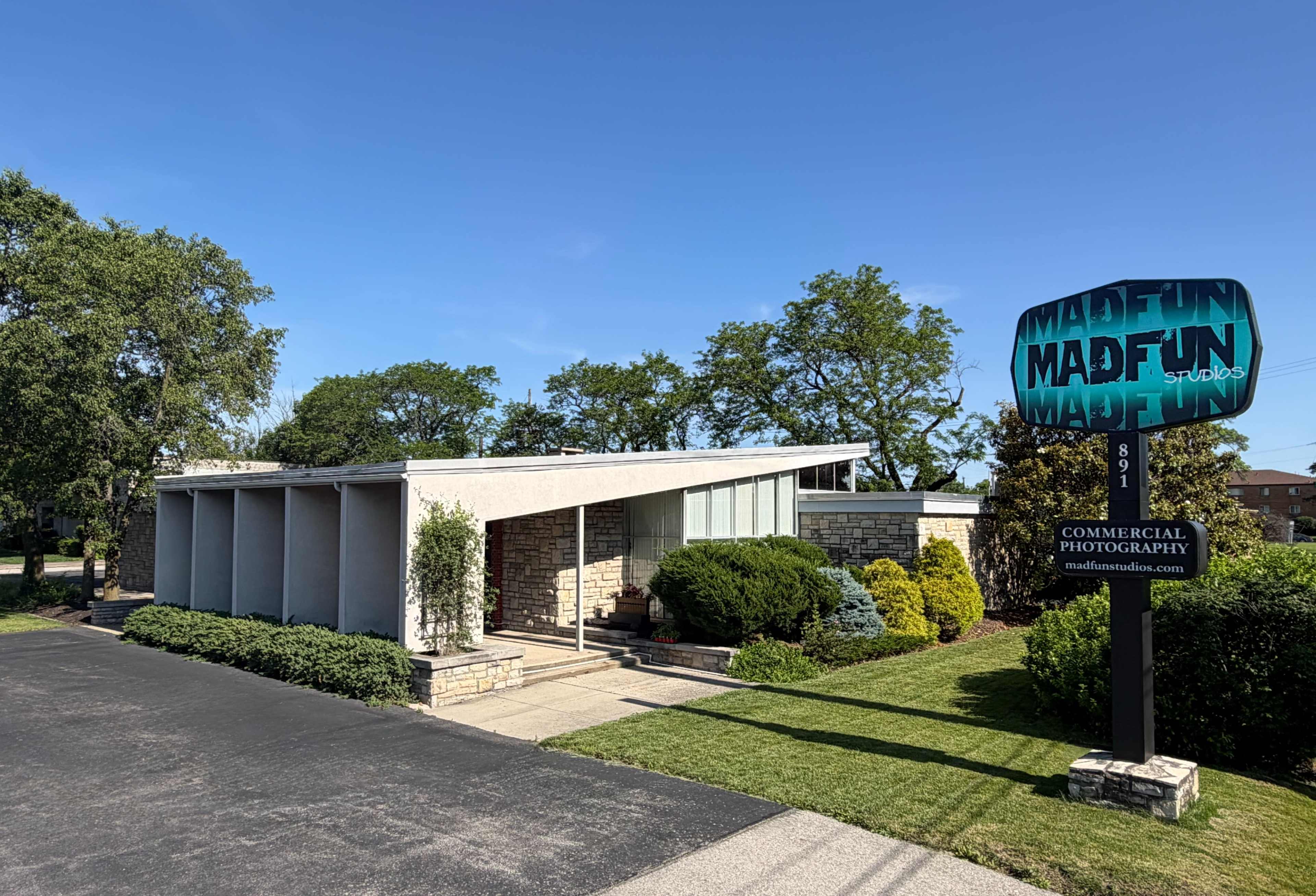 The image shows a modern house with a flat roof, stone façade, and well-manicured landscaping, accompanied by a sign for "MadFun" commercial photography.