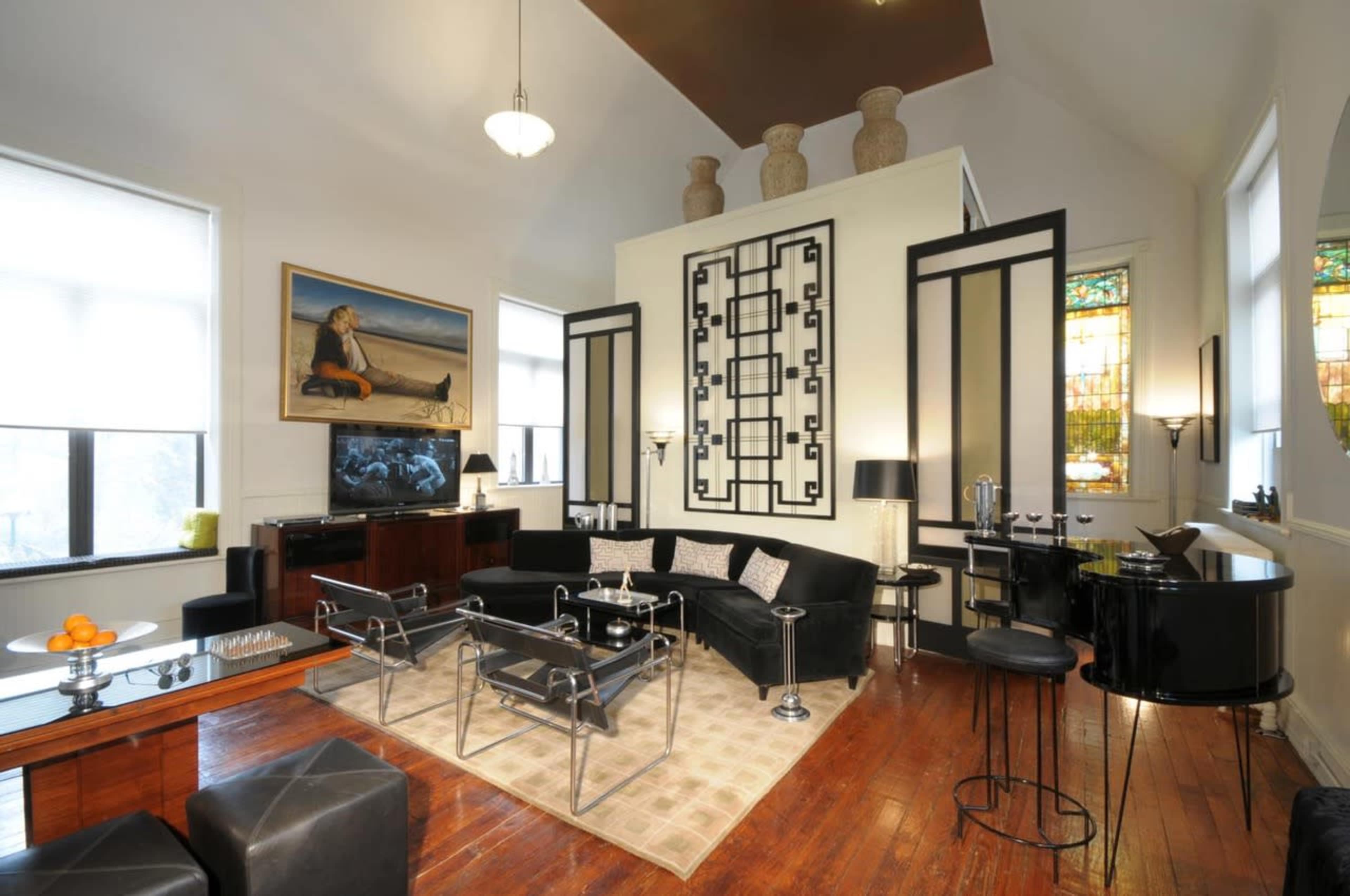 The living room features a high ceiling, large windows, and a modern design with black furniture and a decorative mural.