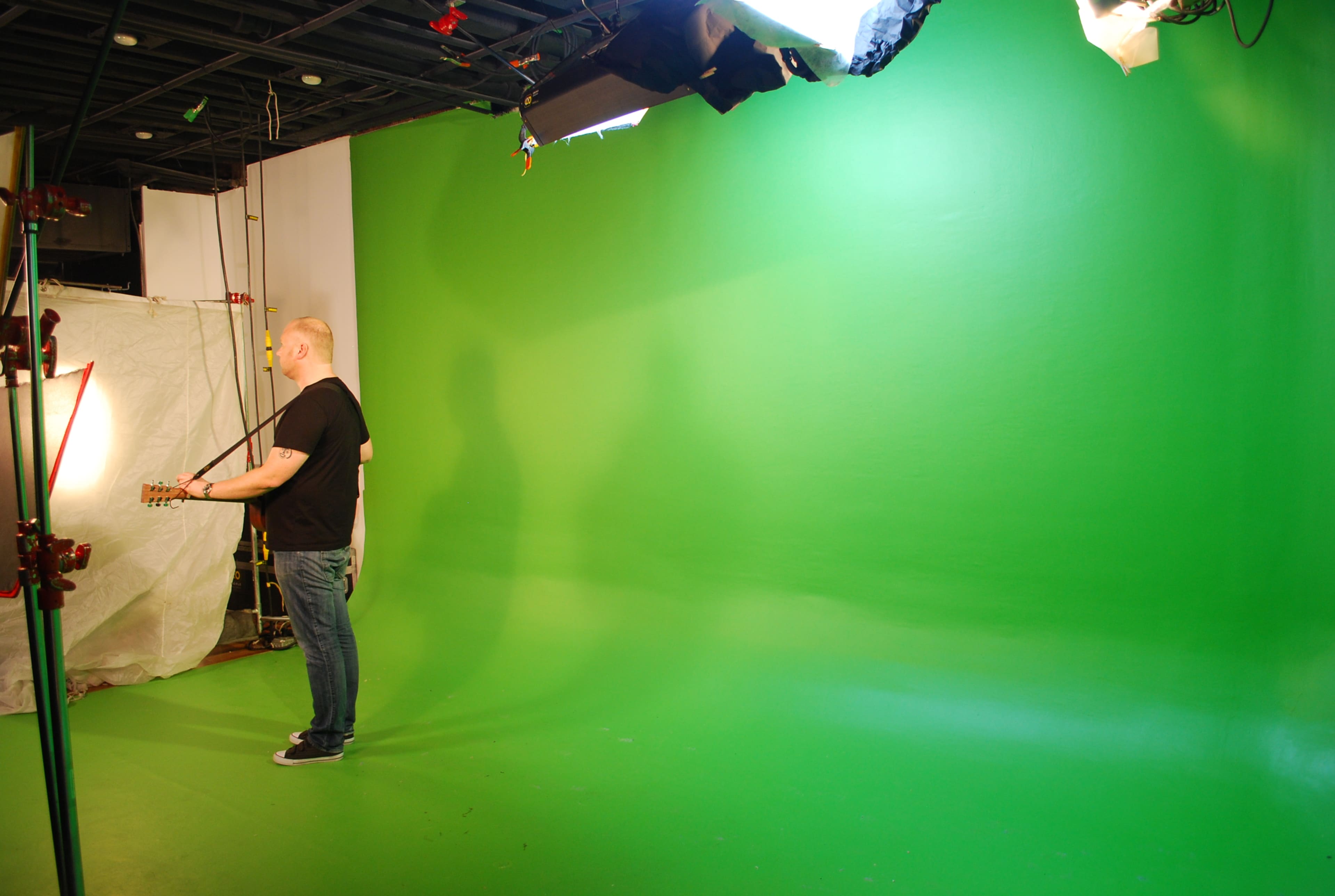 A person stands with a guitar in front of a green screen, with studio lights illuminating the scene.
