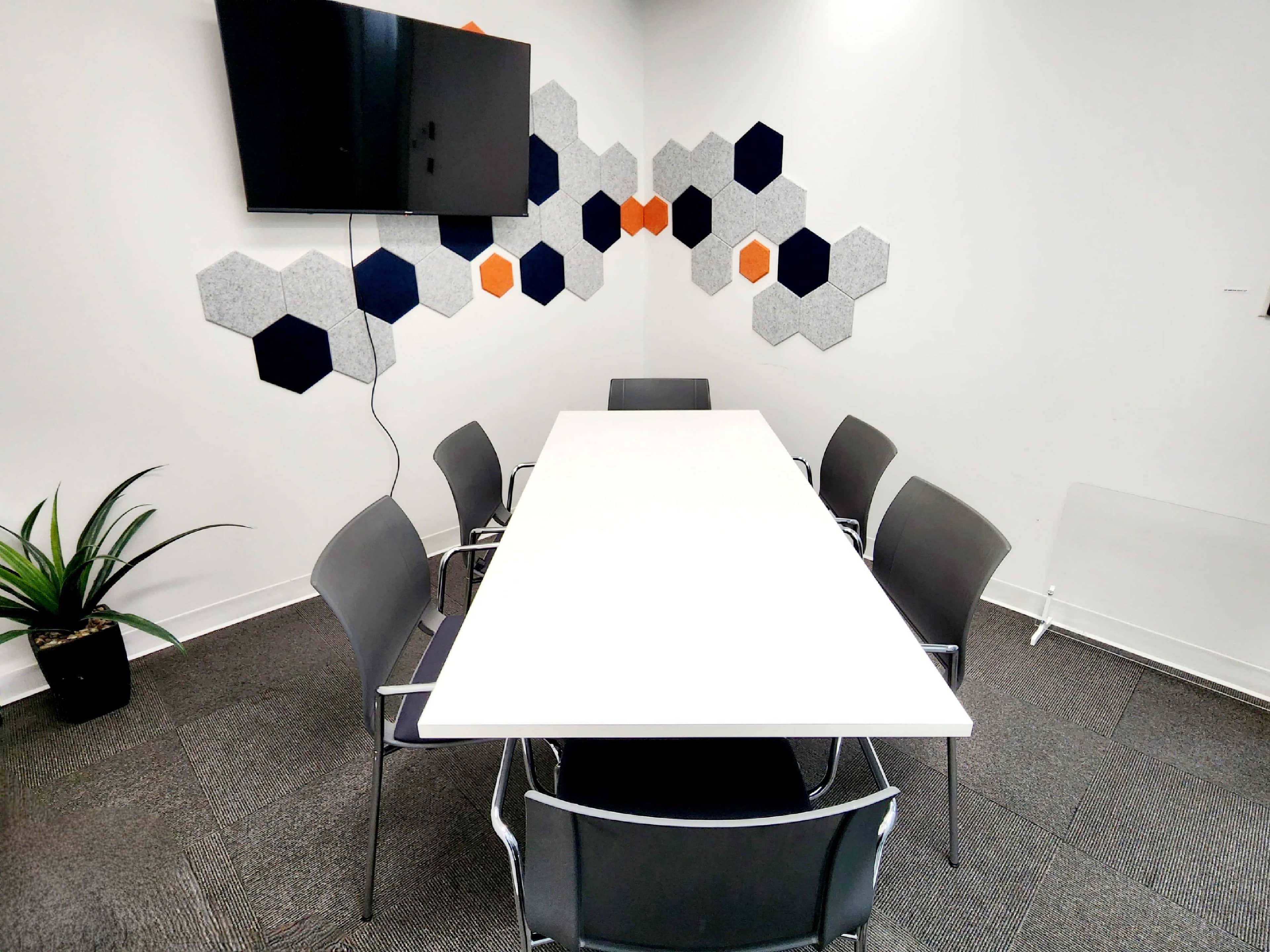 A conference room features a long white table surrounded by six gray chairs, with a television mounted on the wall and hexagonal wall decorations in blue, orange, and gray.