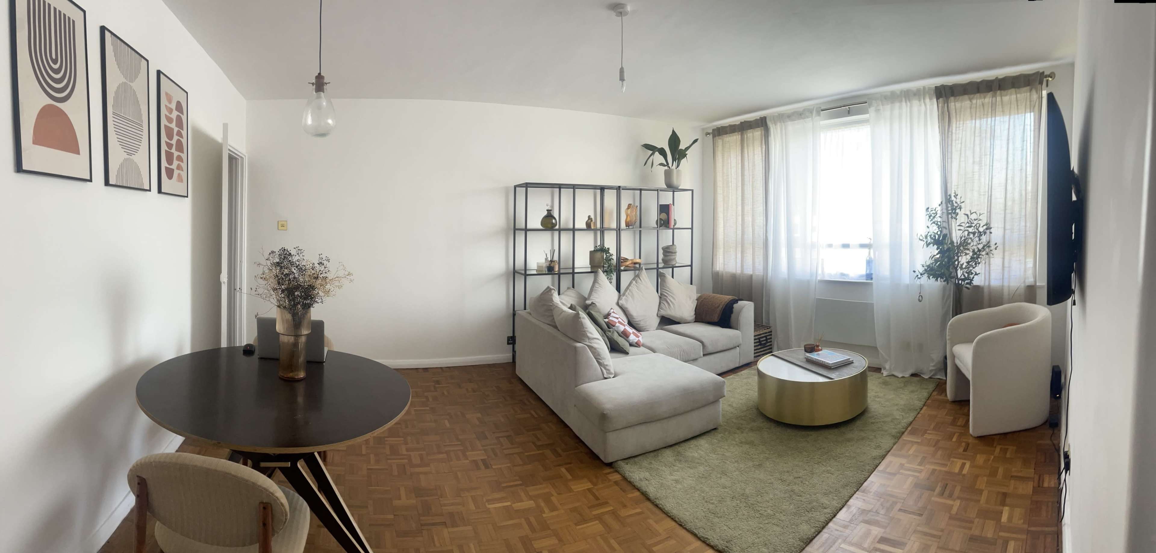 A modern living room featuring a beige sectional sofa, a circular coffee table, a round dining table, and a bookshelf with decorative items, all illuminated by natural light from large windows.