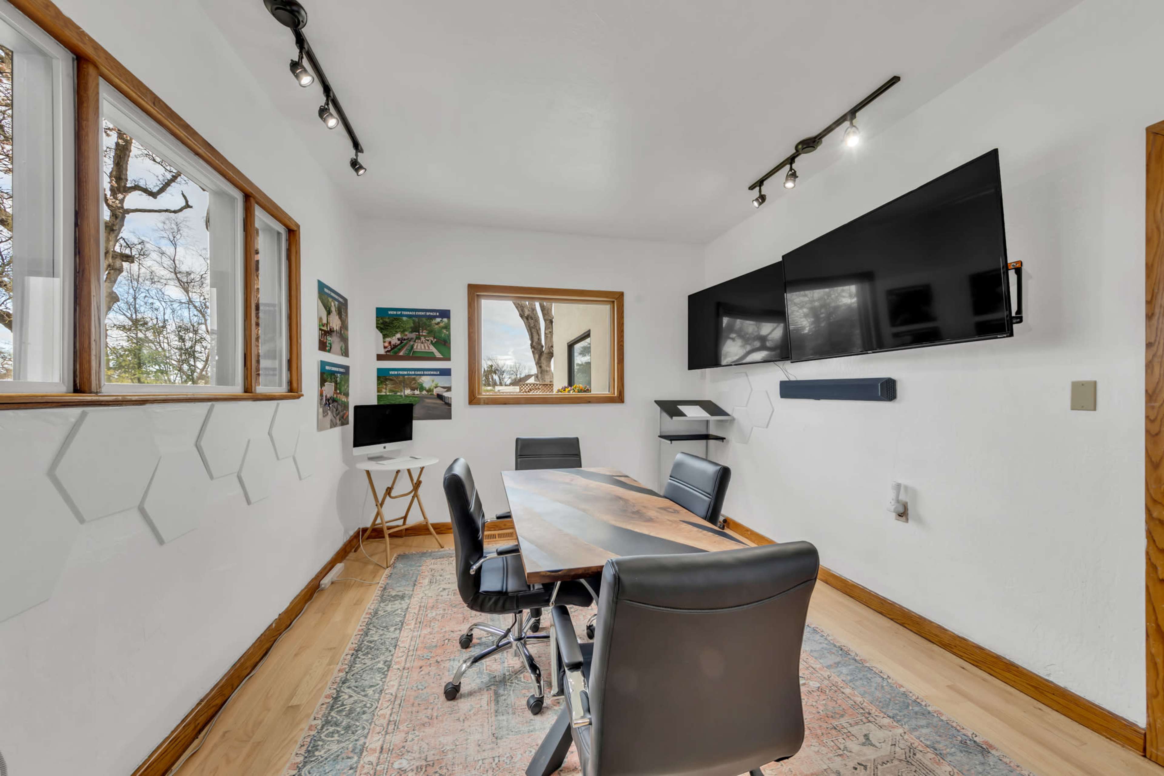 A modern meeting room features a large wooden table surrounded by black office chairs, with dual wall-mounted screens and windows allowing for natural light.