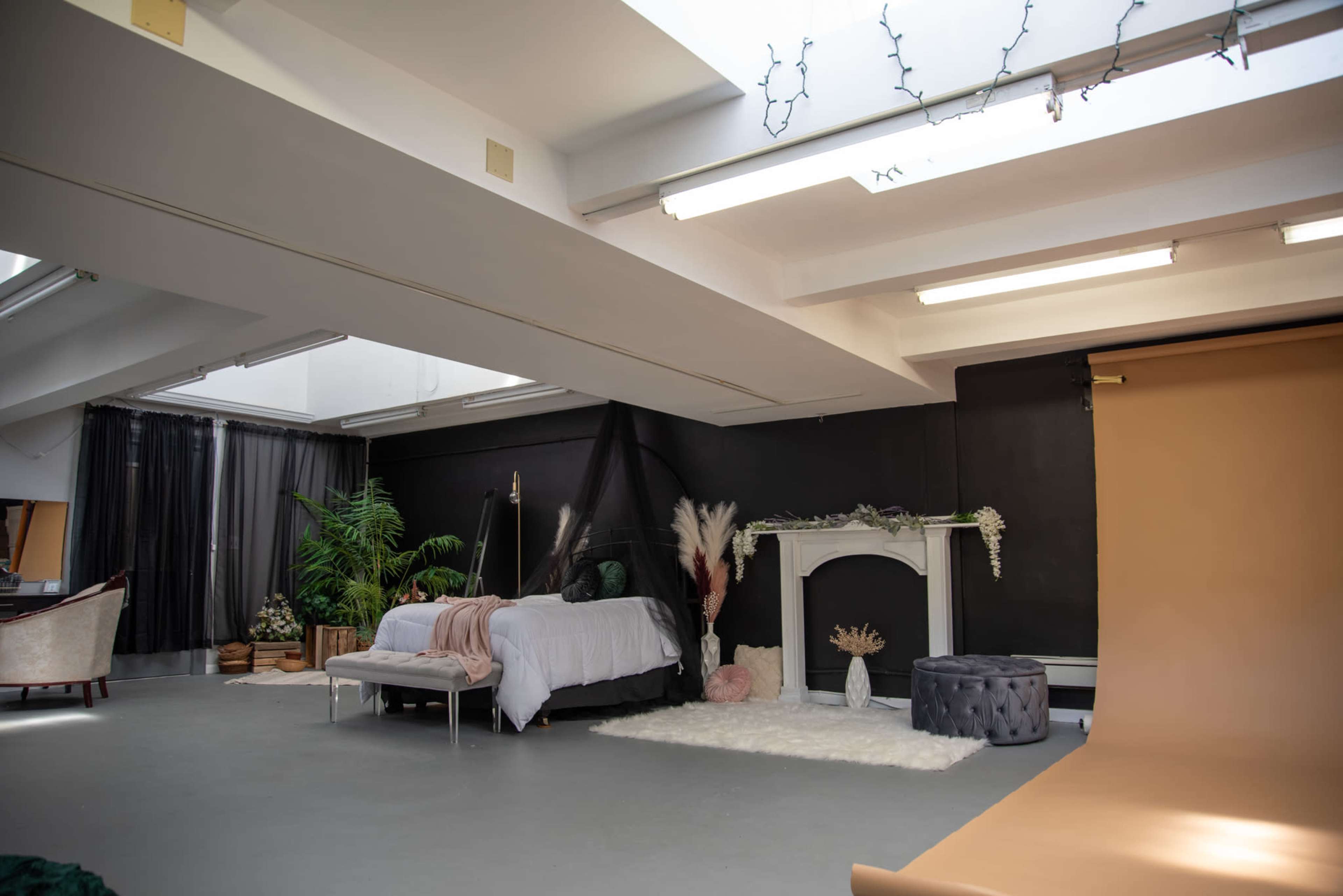 A spacious studio setting with a bed, a decorative fireplace, plants, and a neutral-toned backdrop.
