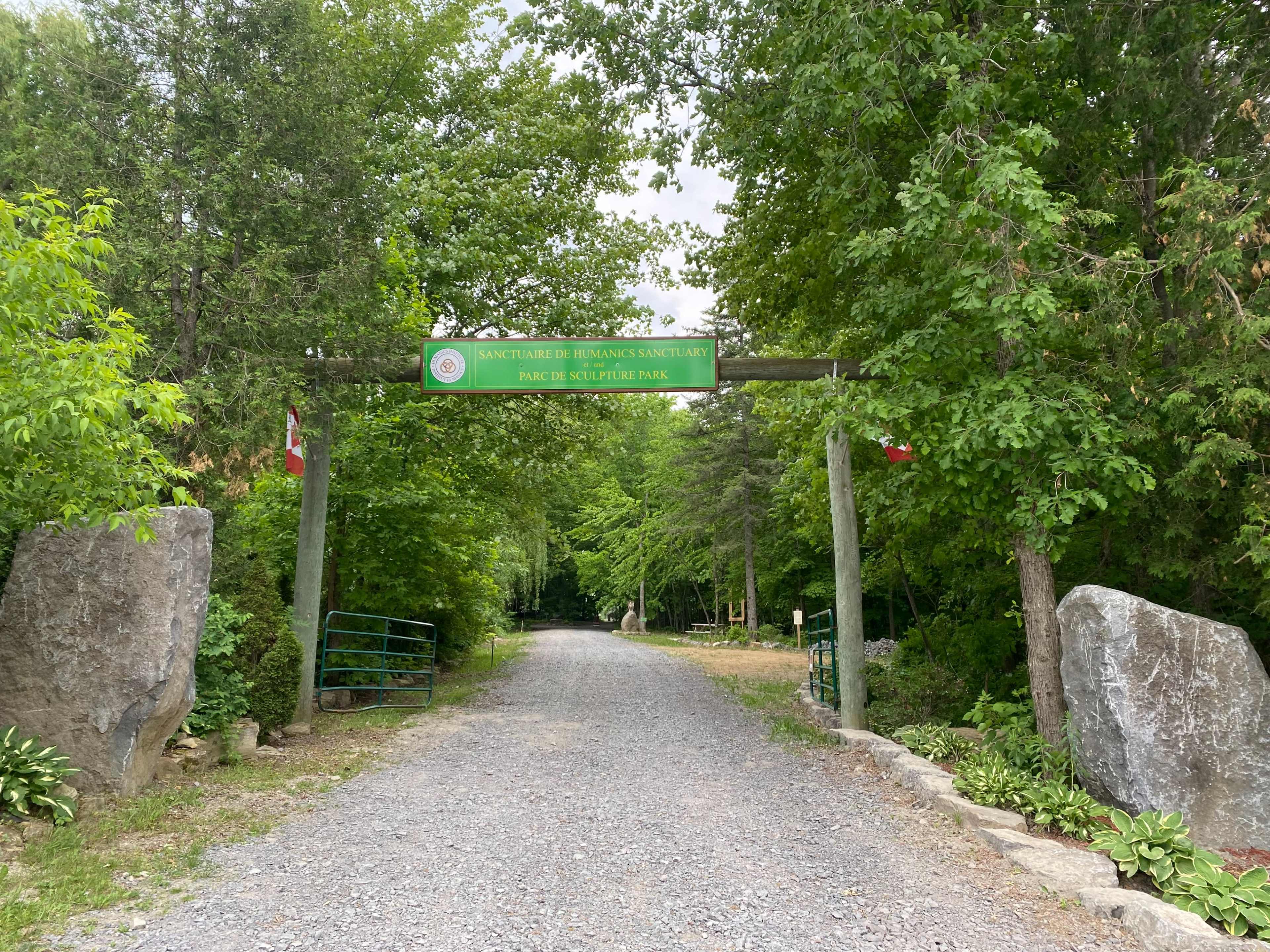 The entrance to a park is marked by a wooden sign and two large stones, surrounded by trees on both sides of a gravel path.